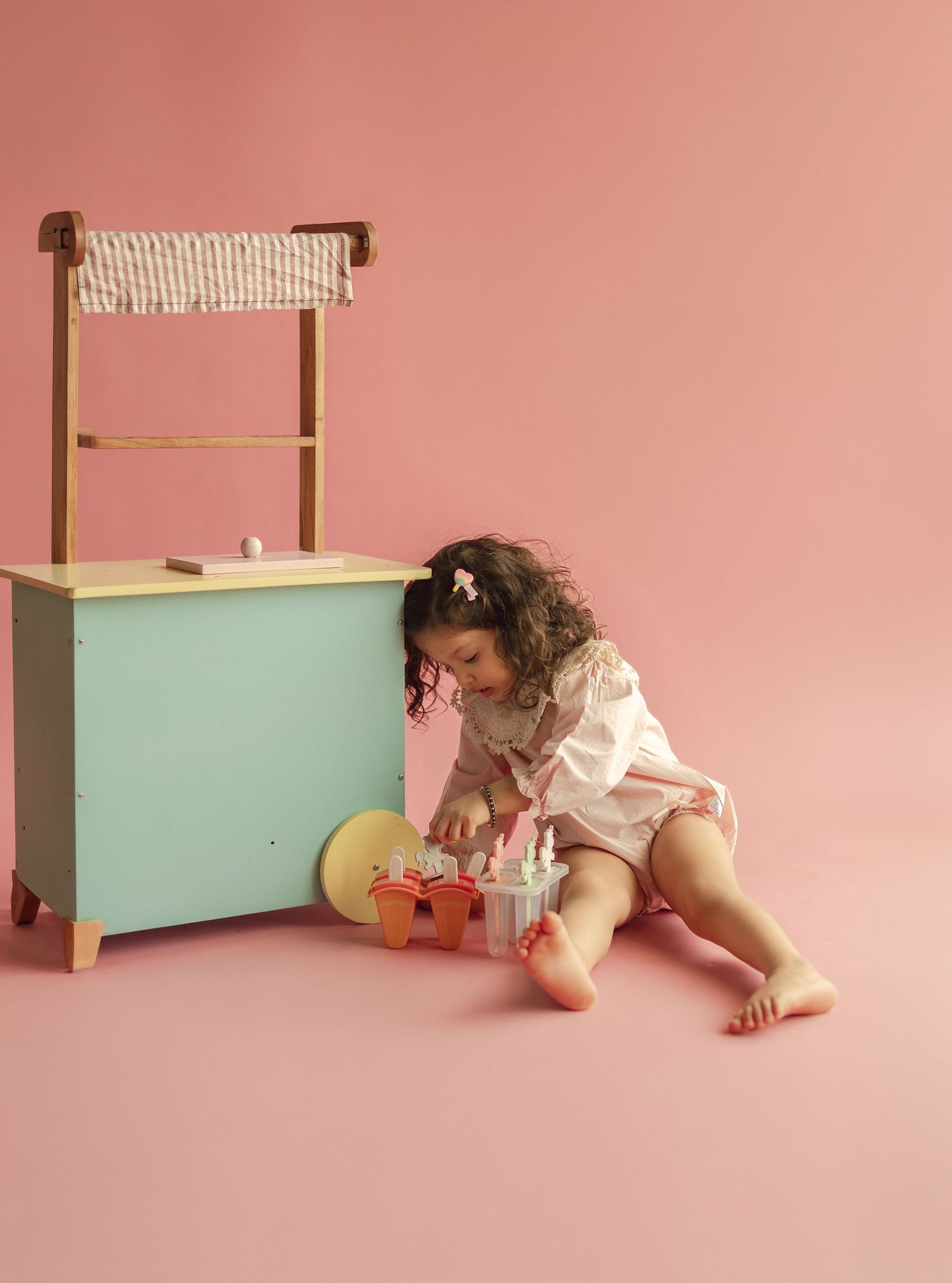 Young toddler girl playing with colorful toy food set beside a mint green wooden play kitchen on pink background.