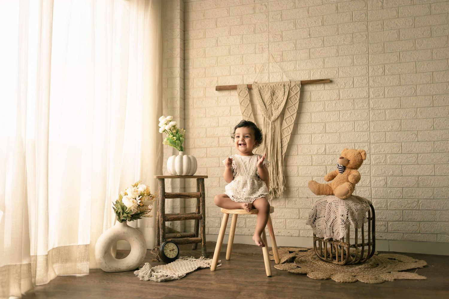 Happy toddler girl sitting on a stool in a boho studio with teddy bear and floral decor.