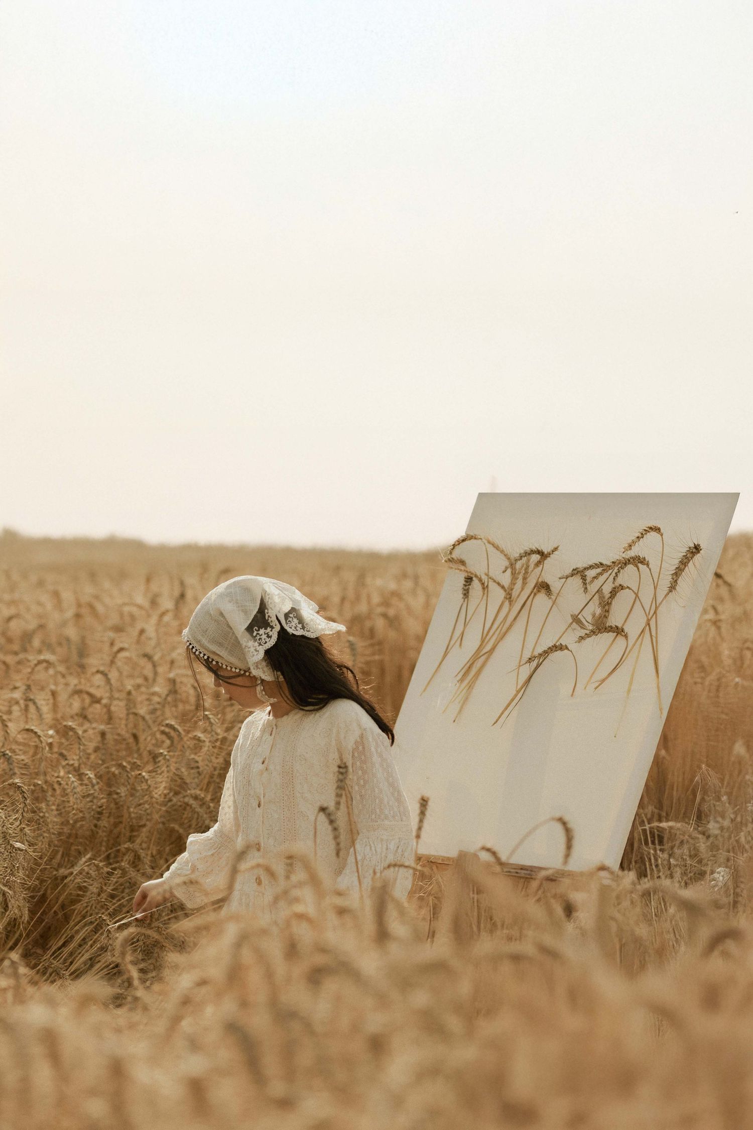 A young girl in a white dress and cap stands in a golden wheat field beside a canvas painting of wheat stalks.
