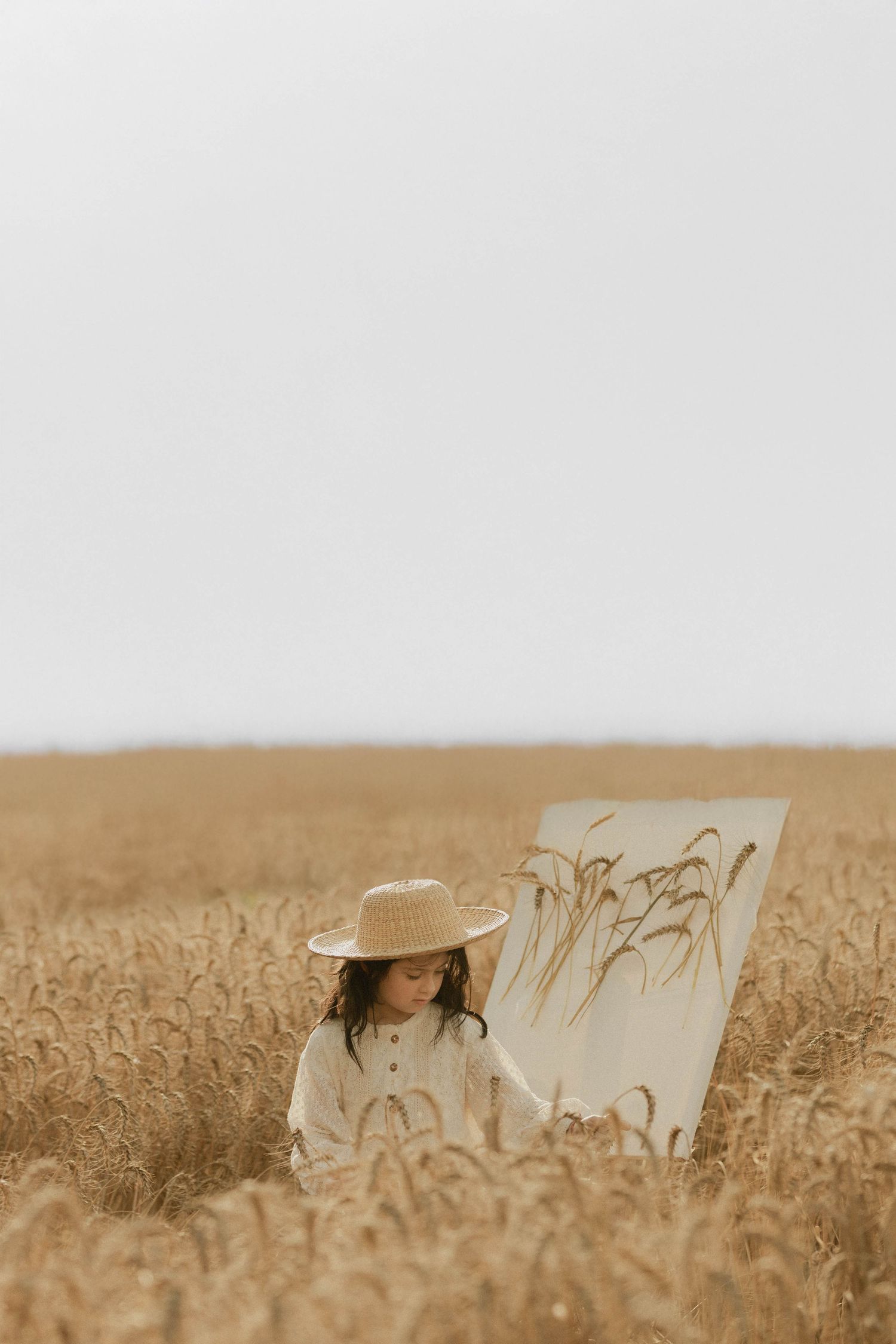 Young girl in straw hat painting wheat stalks on canvas in golden wheat field.