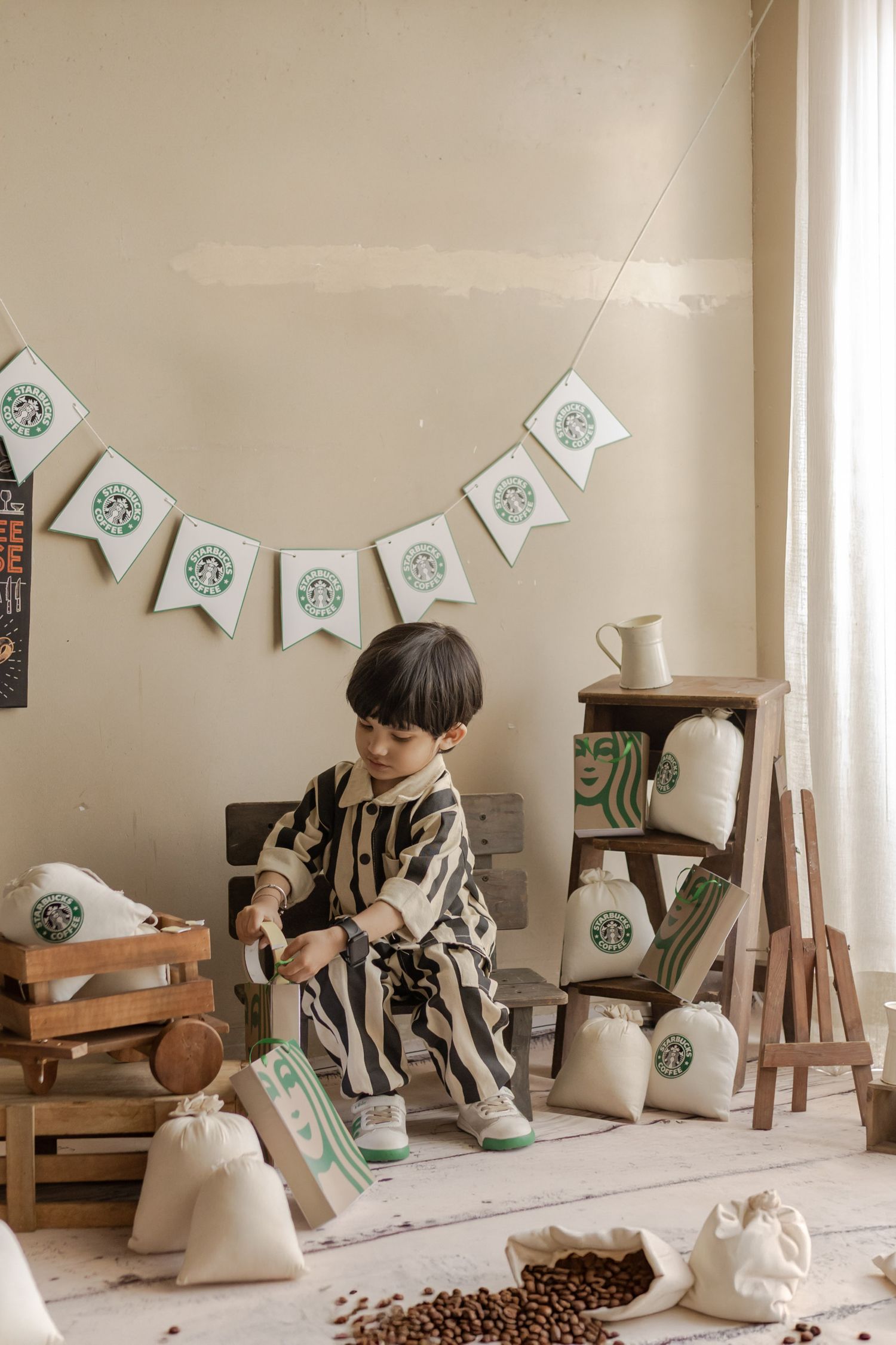 Young boy in striped outfit playing with Starbucks-themed burlap sacks and coffee beans in a decorated room.