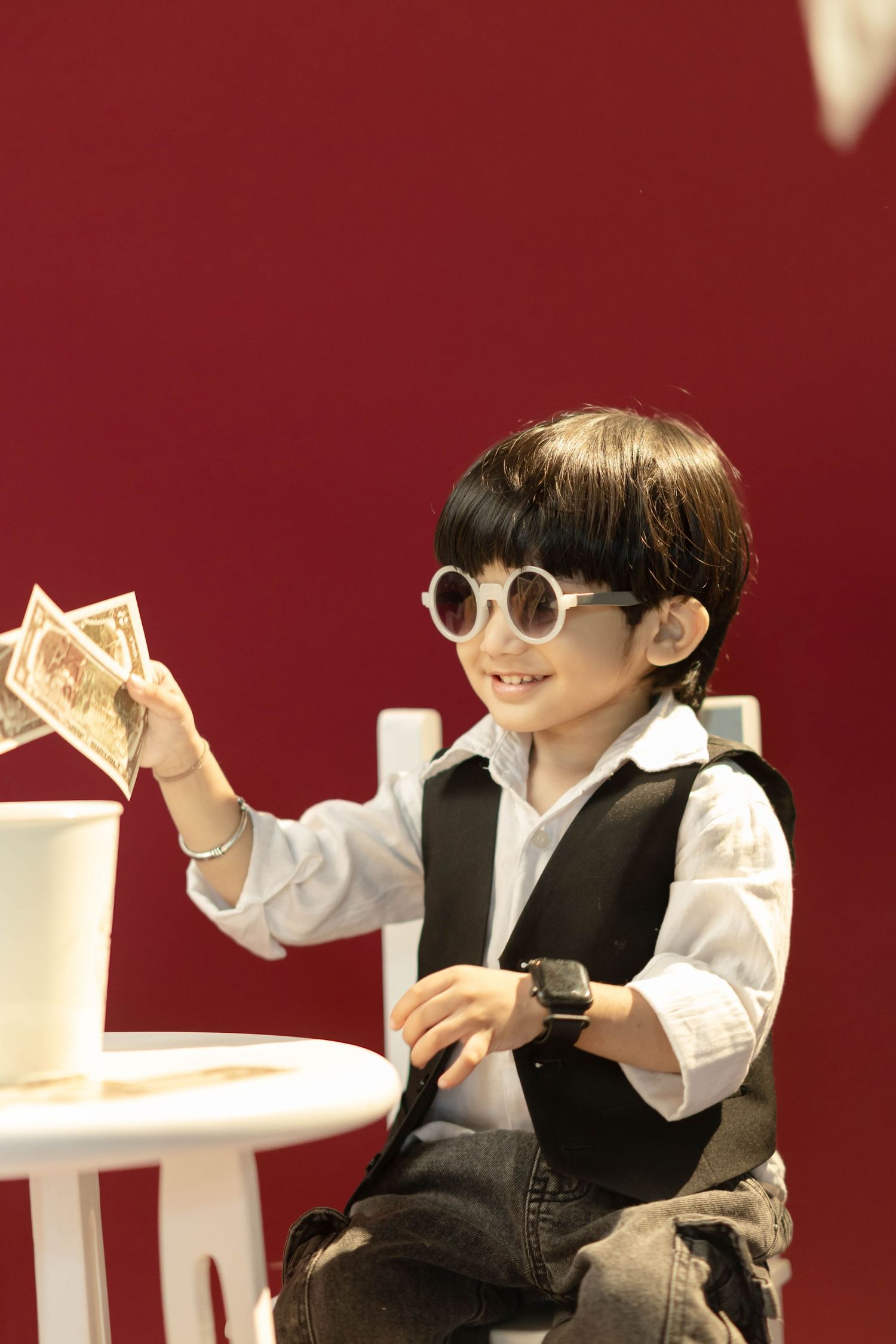 A stylish young boy wearing round white sunglasses and a vest holds money while sitting at a white table against a red background.