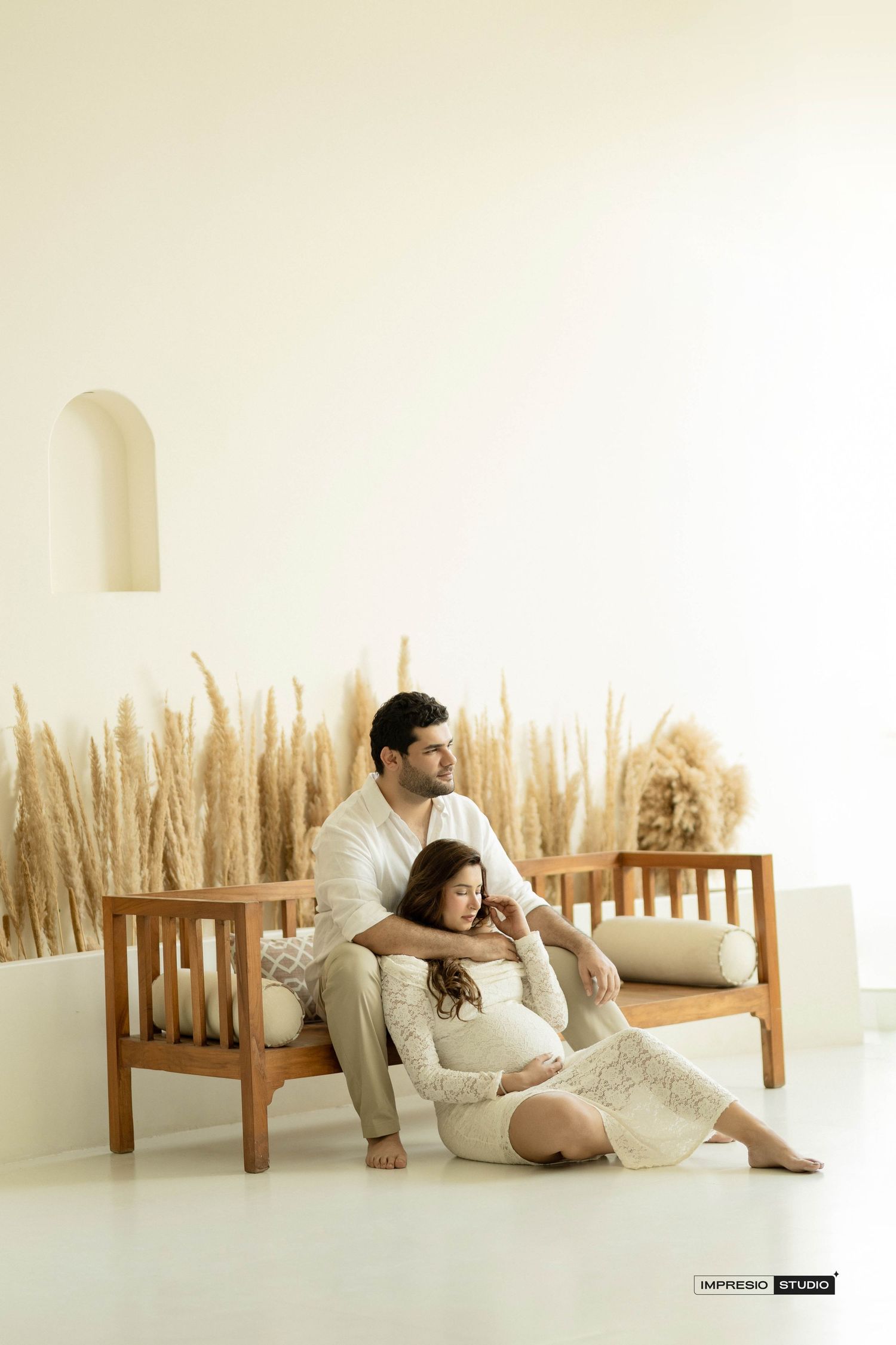 Couple in white outfits posing on wooden bench with dried pampas grass backdrop in a bright studio.