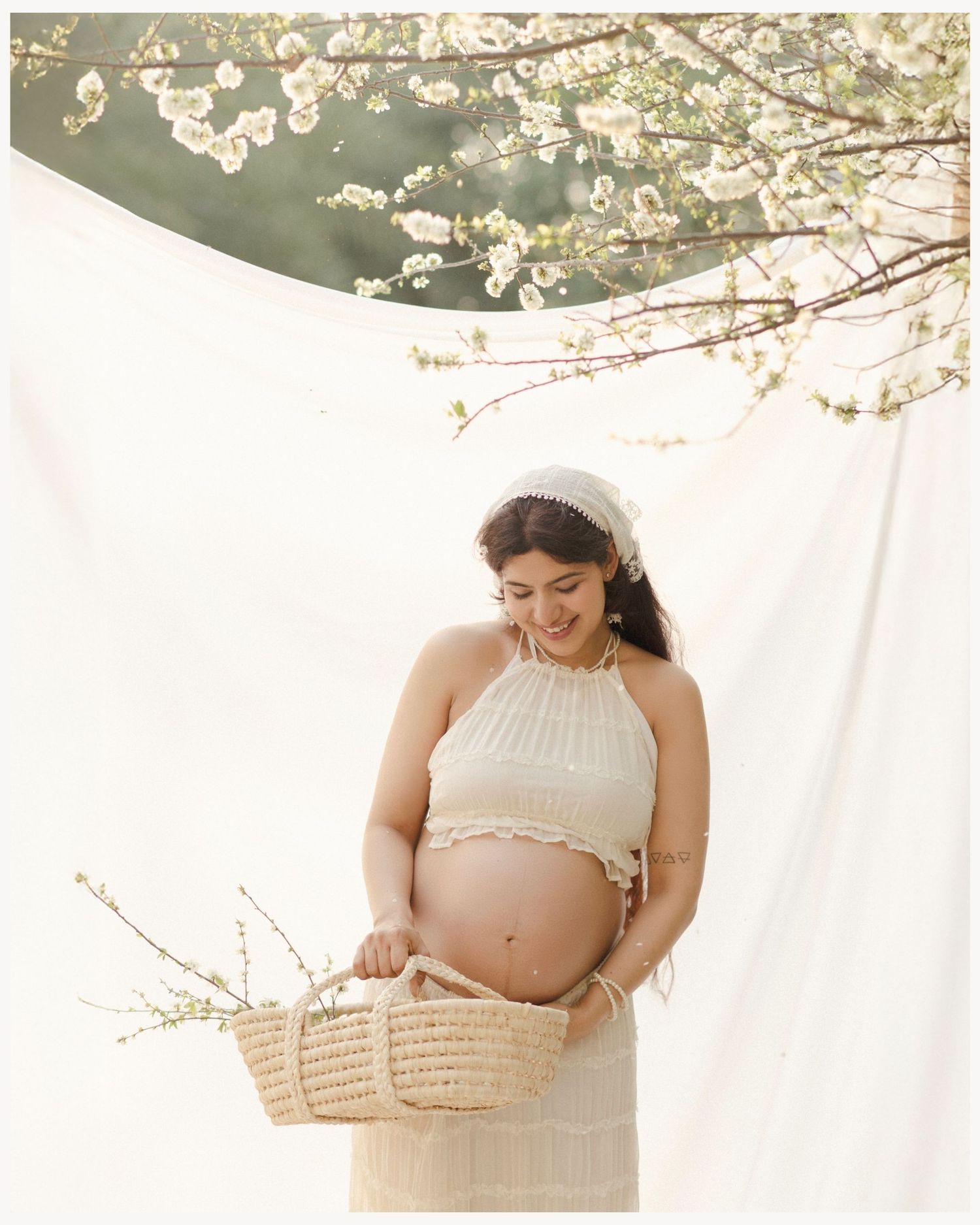 Pregnant woman in cream halter top holding wicker basket under blooming cherry blossom tree with white fabric backdrop.