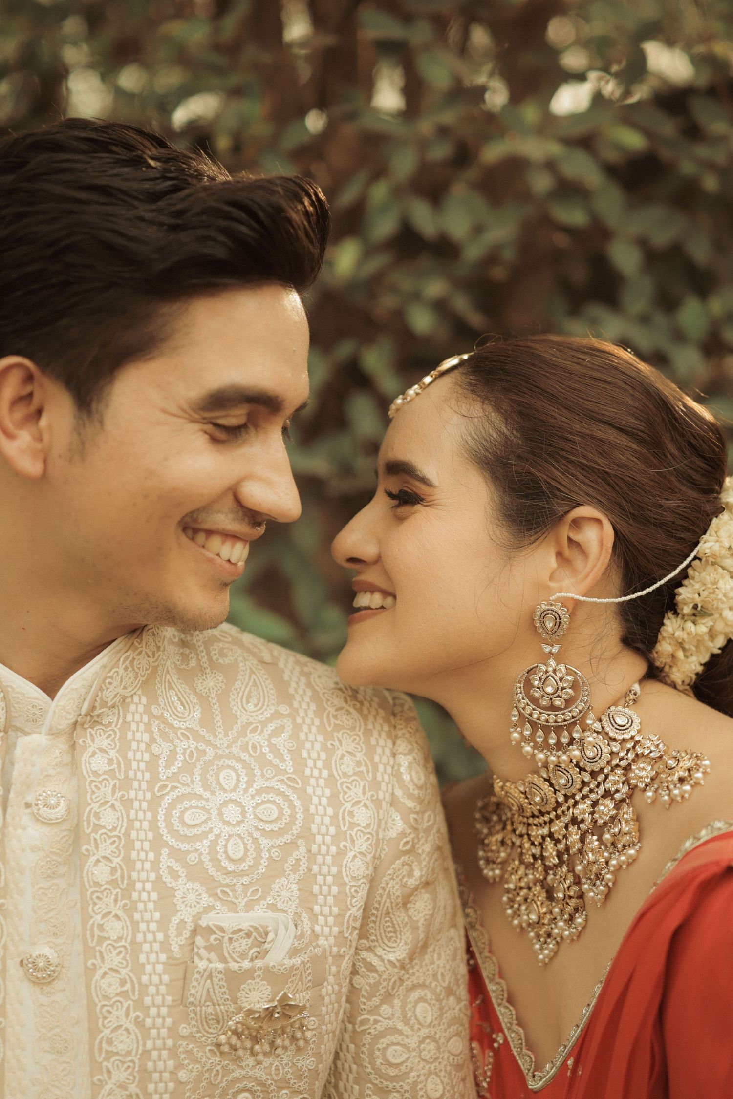 A smiling couple in traditional Indian wedding attire, man in cream sherwani, woman in red with gold jewelry, facing each other.