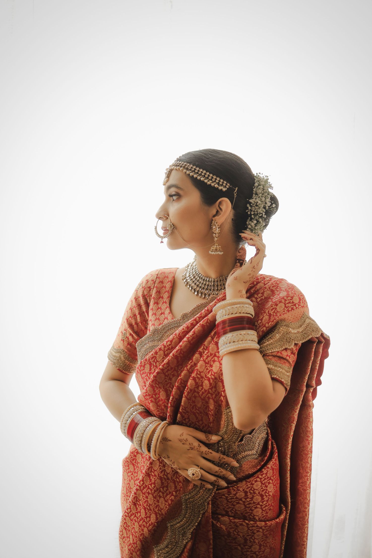 Woman in red saree with traditional Indian bridal jewelry, maang tikka, nath, and bangles posing gracefully.
