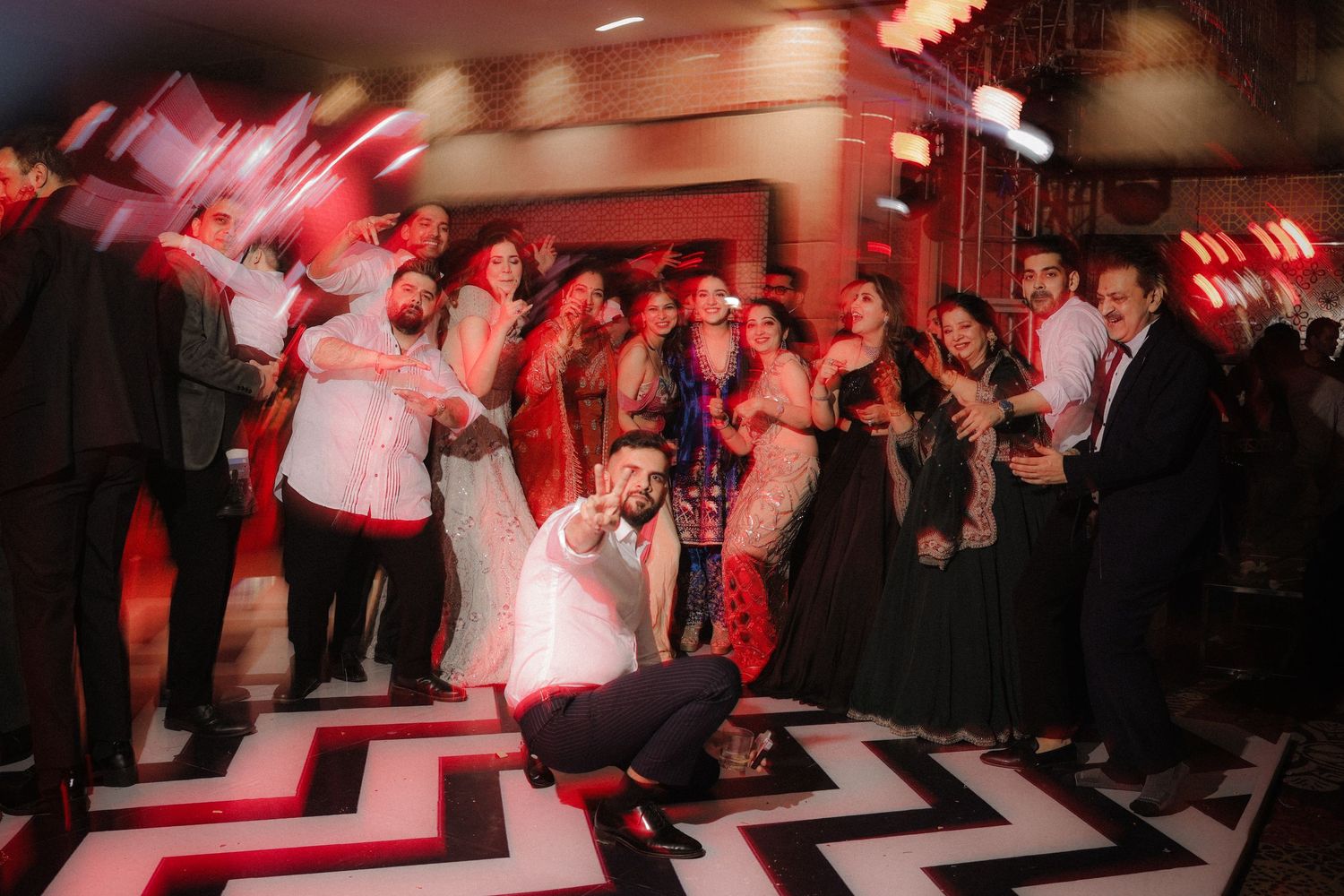 People dancing and celebrating on a black and white chevron patterned dance floor under red mood lighting at a party.