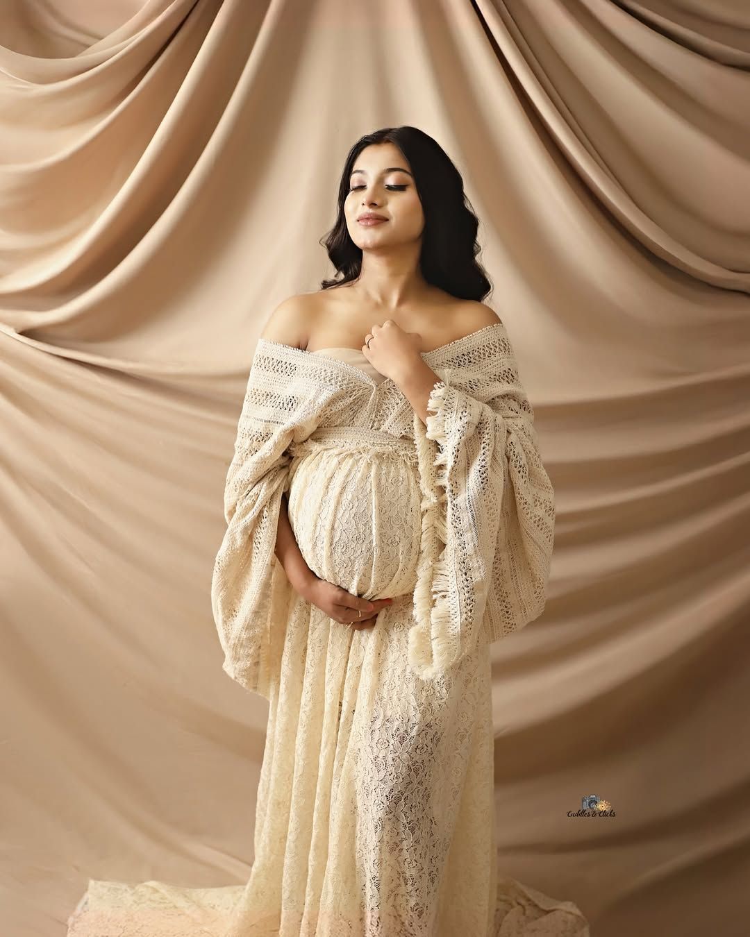 Elegant maternity photo with cream lace dress against draped fabric backdrop.