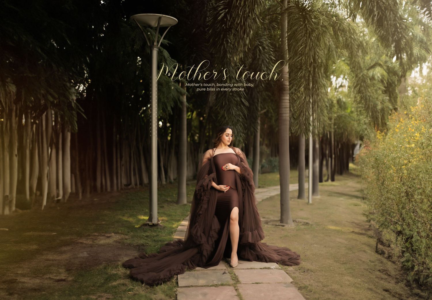 Outdoor maternity photo session with long brown dress on tree-lined path with street lamps.