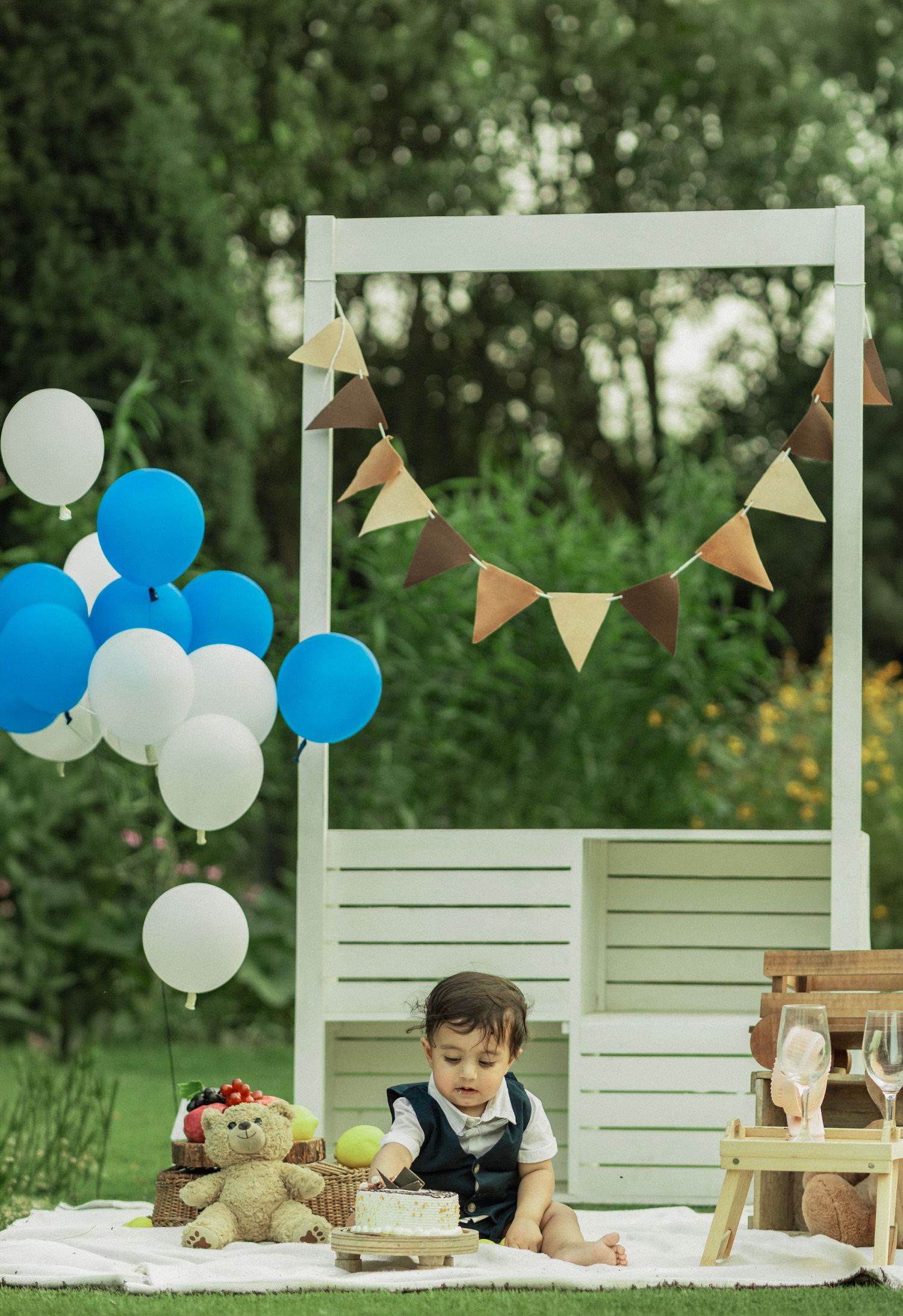 Baby boy sits outdoors at first birthday party with blue and white balloons, bunting, teddy bear, and cake on white blanket.