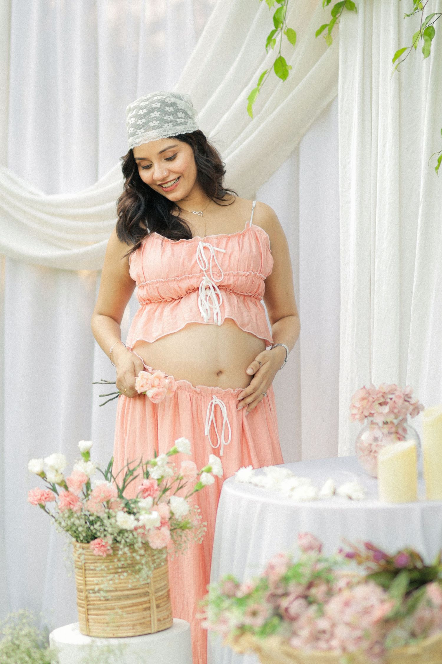 Smiling pregnant woman in pink crop top and skirt cradling baby bump at floral baby shower.