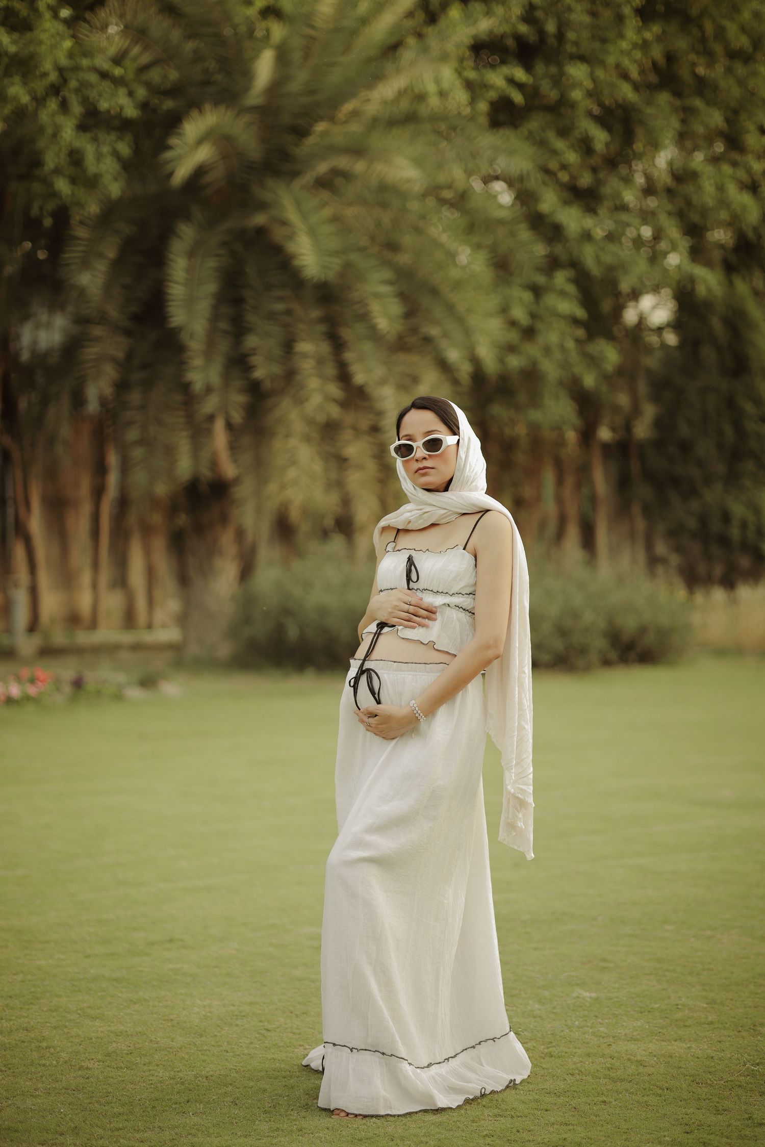 Pregnant woman in white maxi dress and head scarf poses outdoors on green lawn with tropical trees.