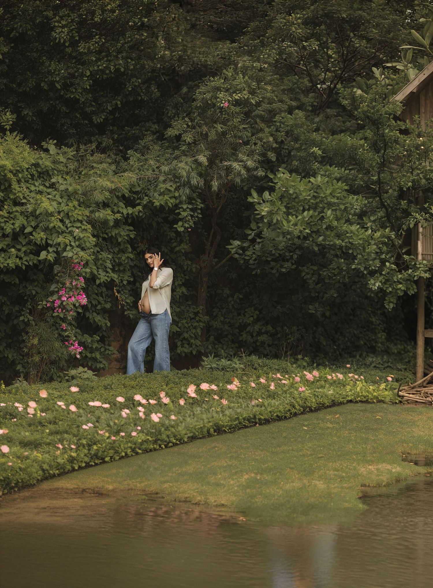 A couple embraces near a serene pond bordered by pink flowers, surrounded by lush green trees.