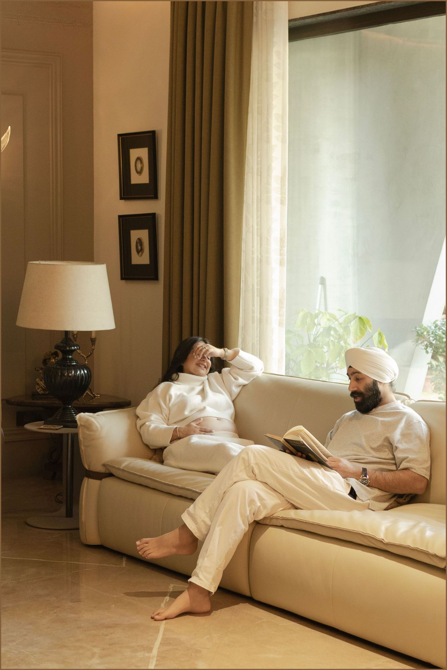 A Sikh man in a white turban reads a book on a cream sofa beside a relaxed woman in a bright living room.