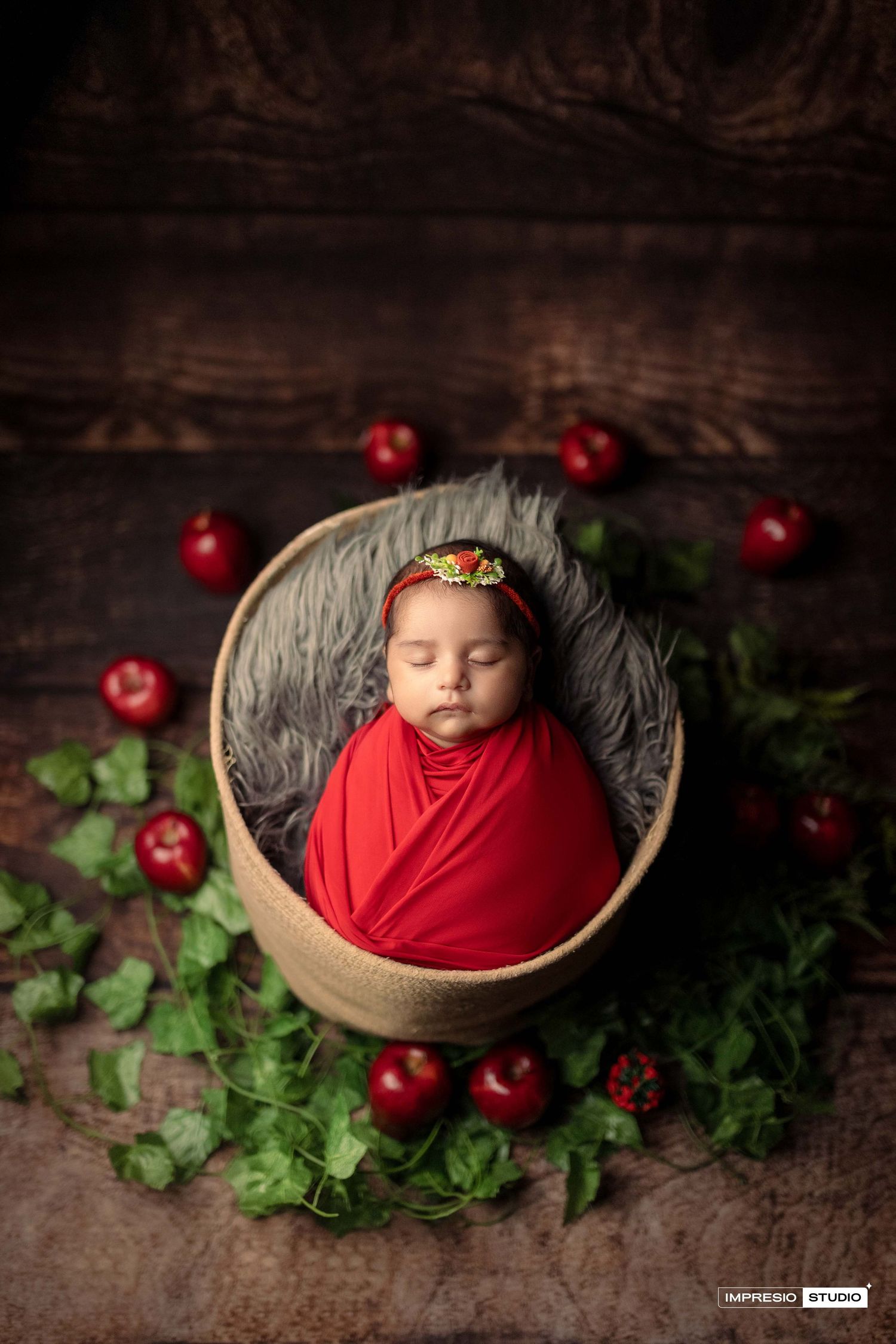 Newborn baby swaddled in red wrap sleeping in a wooden bowl basket surrounded by red apples and green leaves.
