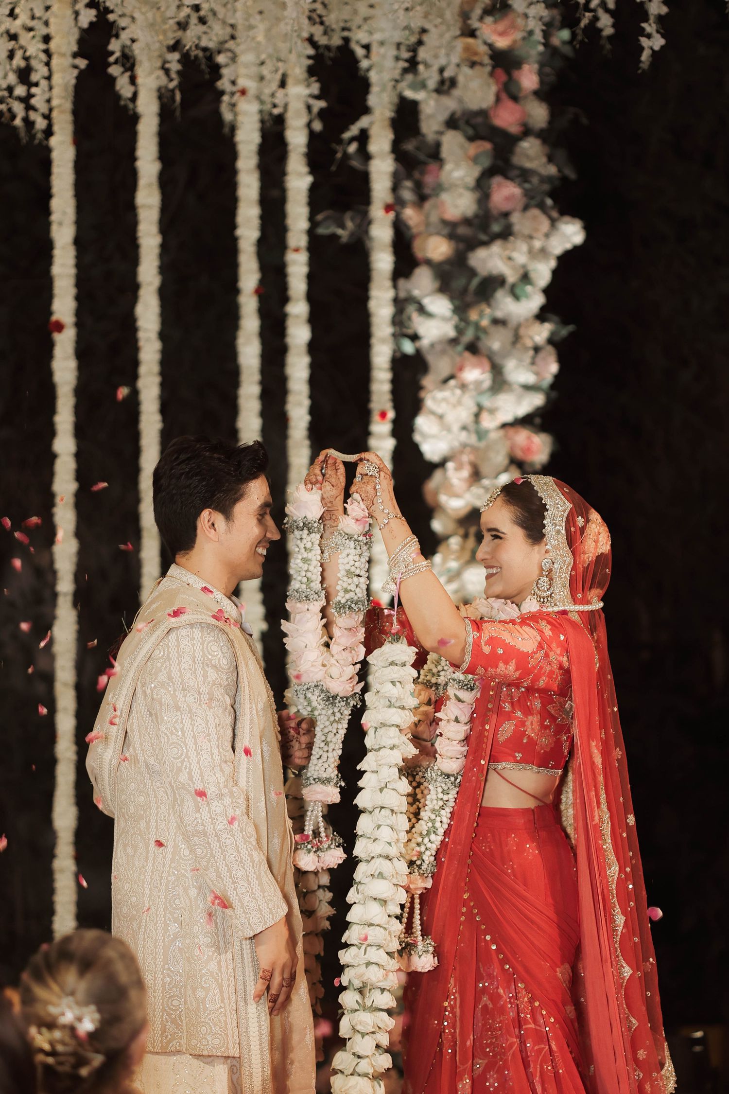 Couple in traditional Indian wedding attire exchanging garlands during a jaimala ceremony with floral backdrop.
