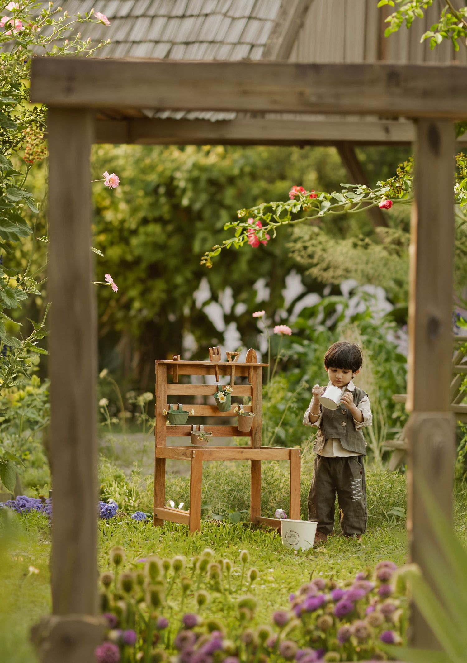Young boy playing in a lush garden surrounded by wooden furniture, colorful flowers, and greenery on a sunny day.
