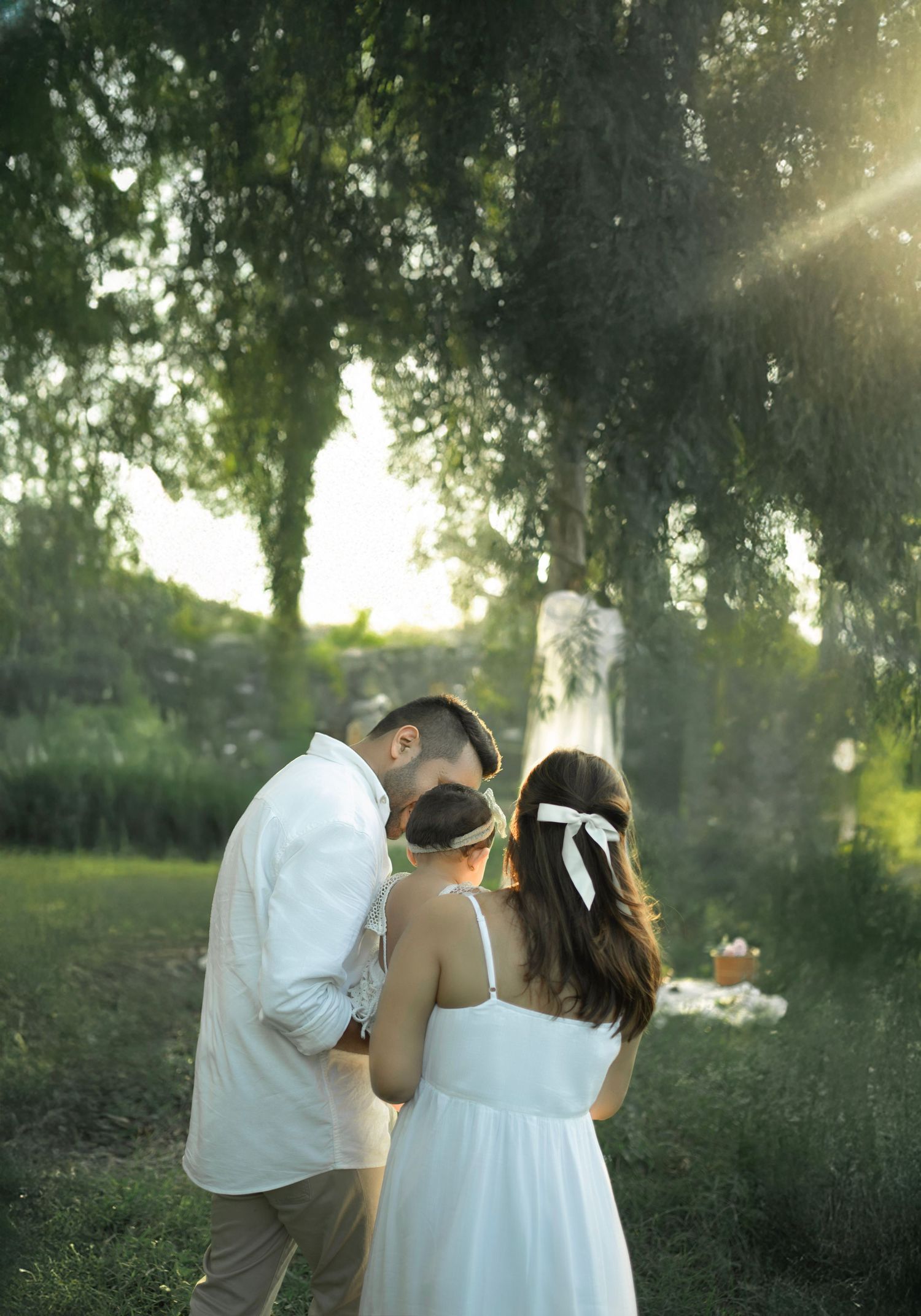A couple and child share a tender moment in a sunlit garden, dressed in white, surrounded by lush green trees.