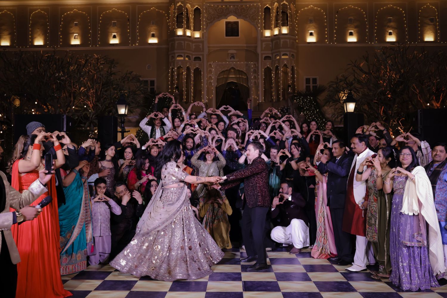 Elegant dancers in formal attire perform on a checkered ballroom floor at an ornate venue with illuminated walls.