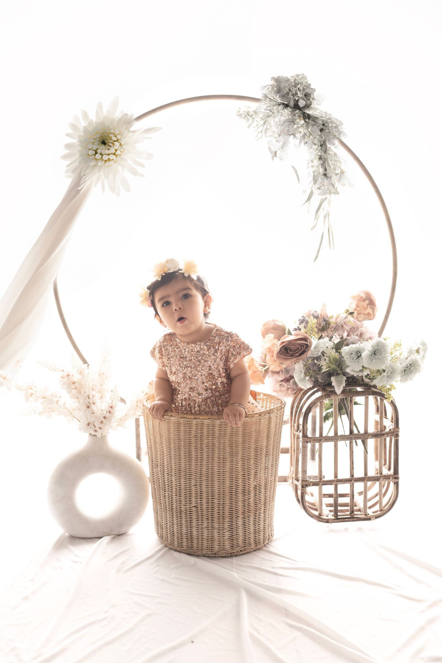 Baby girl in sequin dress sits in wicker basket surrounded by floral decor and circular arch in bright studio setting.