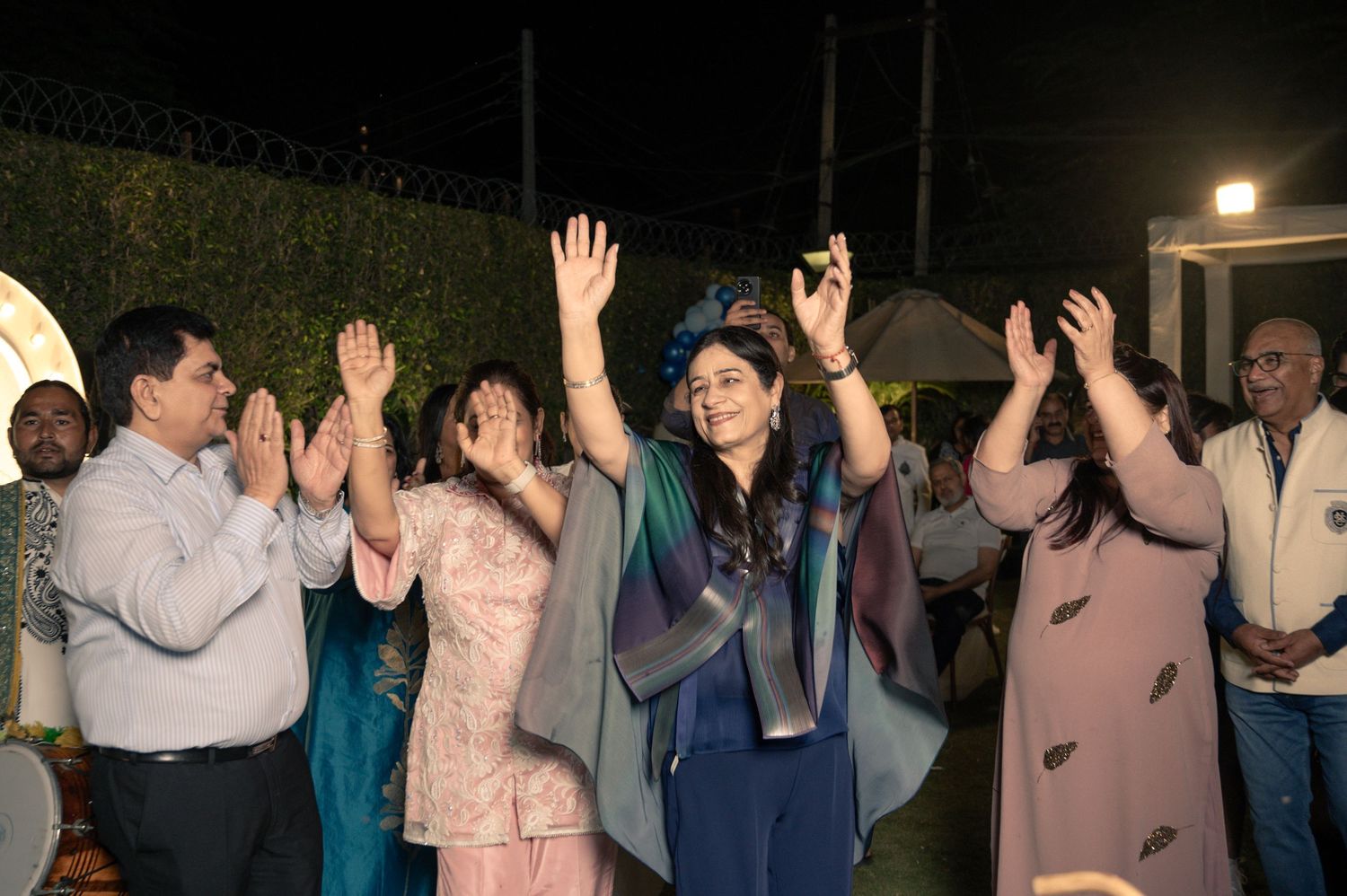 Group of people dancing and celebrating at an outdoor evening party, woman in blue outfit raises hands joyfully.