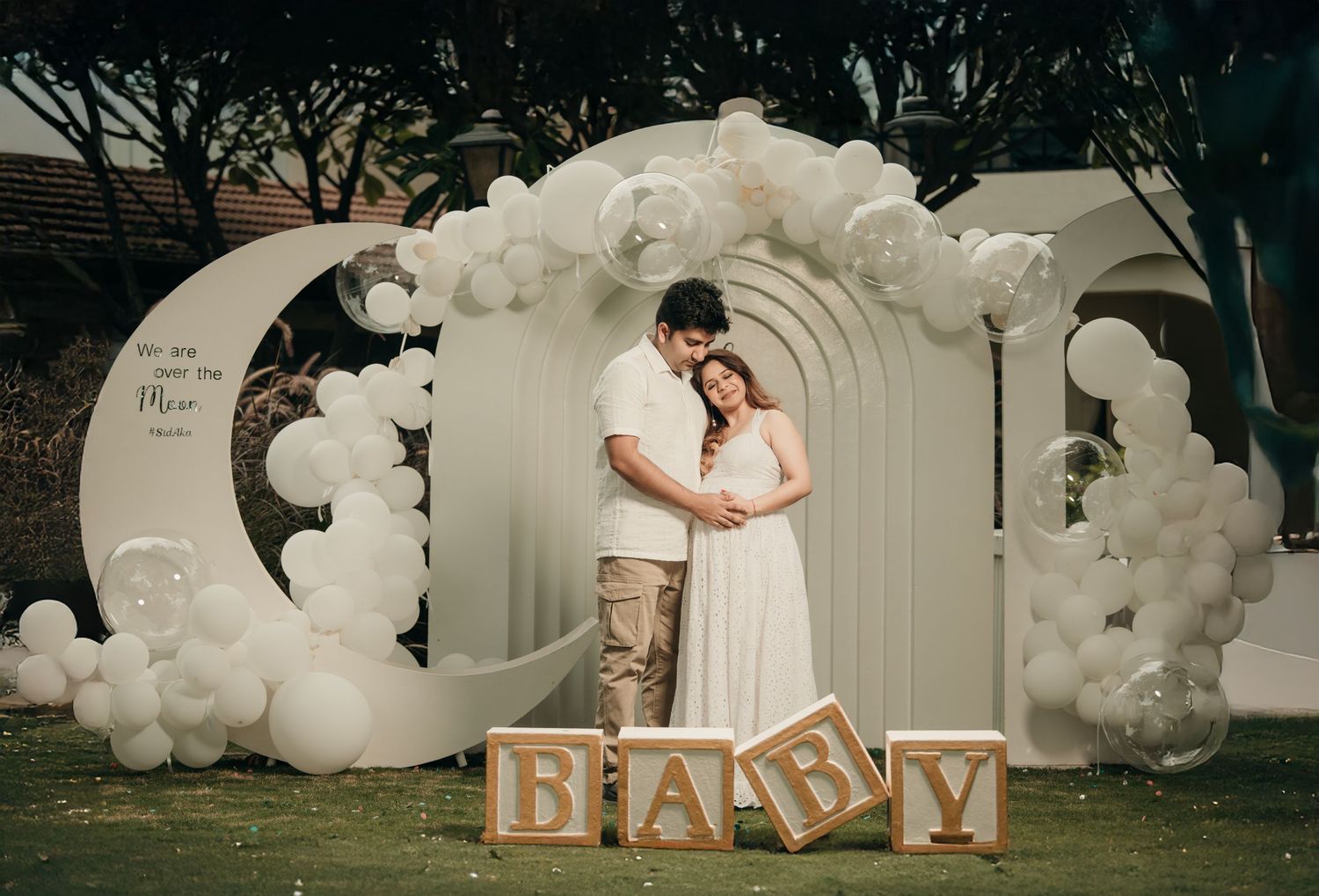 Couple embracing at elegant baby shower with white balloon arch and wooden BABY blocks on green lawn.
