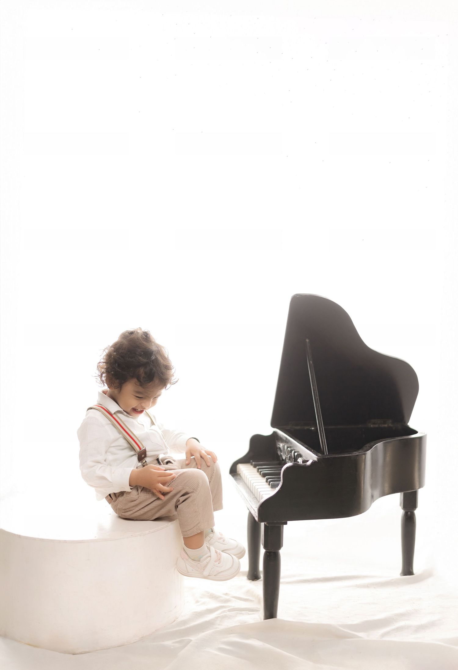 Young child in suspenders sitting beside a small black grand piano on a white background.