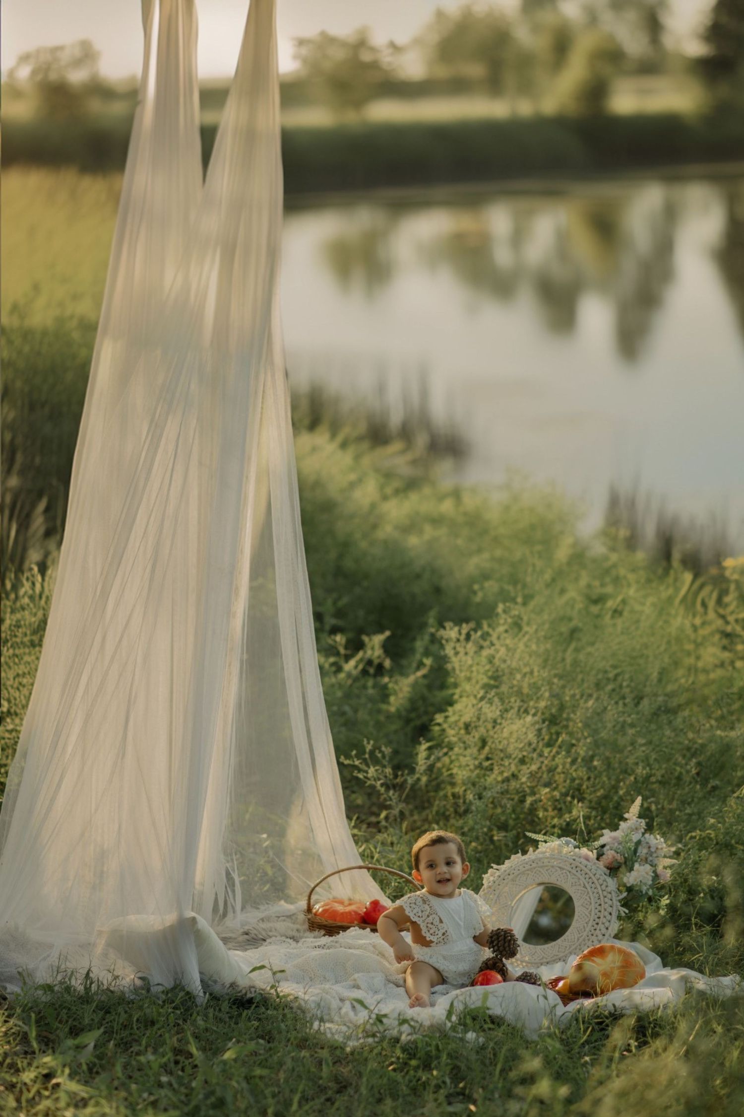 Baby girl in white dress sits on blanket by serene lake with flowing white canopy and toys at golden hour.