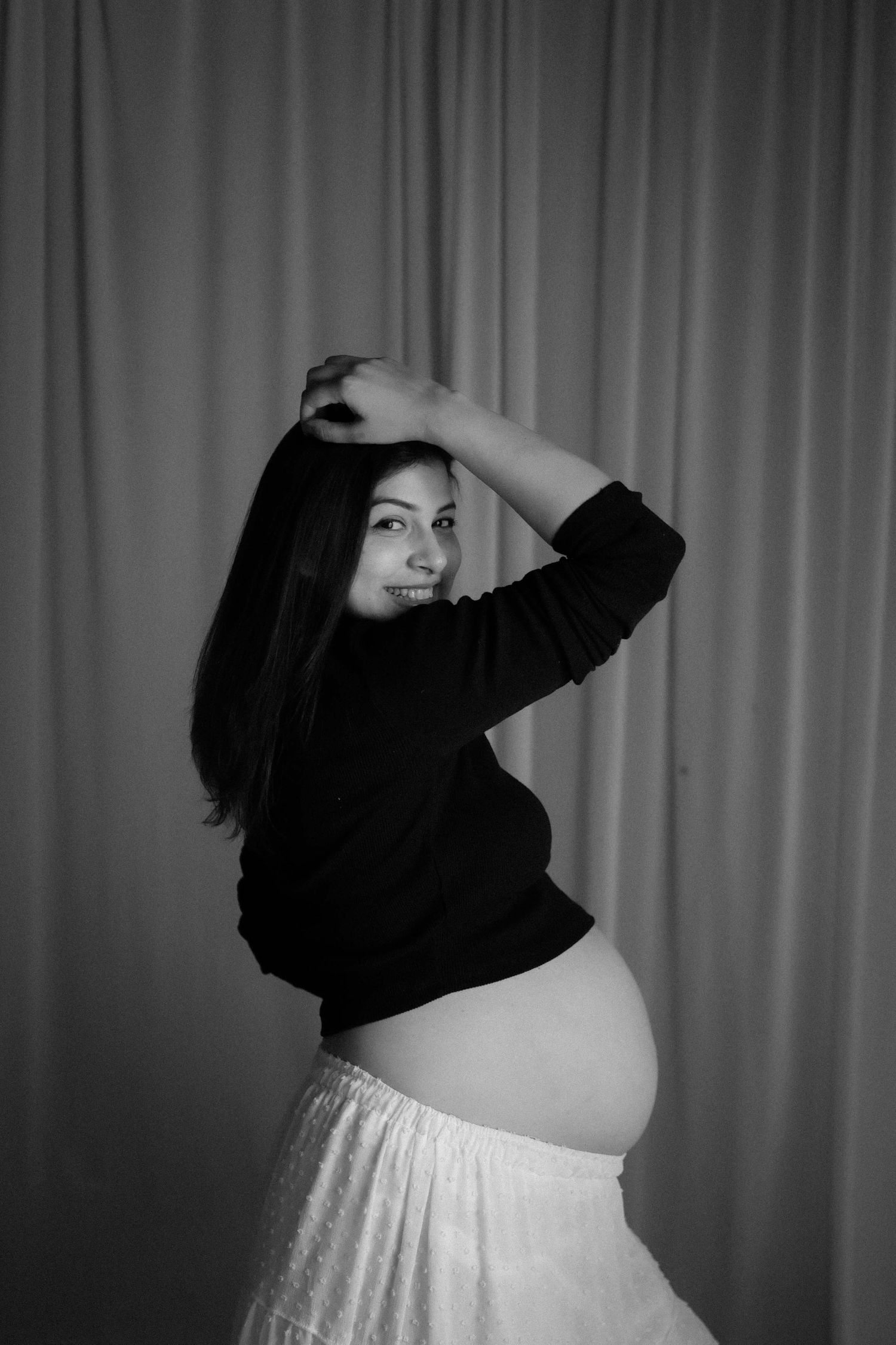 Black and white photo of a smiling pregnant woman in a black top and white skirt, posing with hands raised near her head.