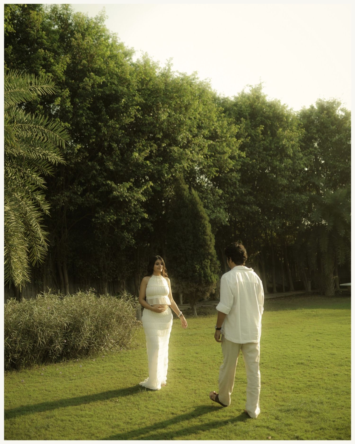 Pregnant woman in white dress with partner strolling on lush green lawn surrounded by tall trees.
