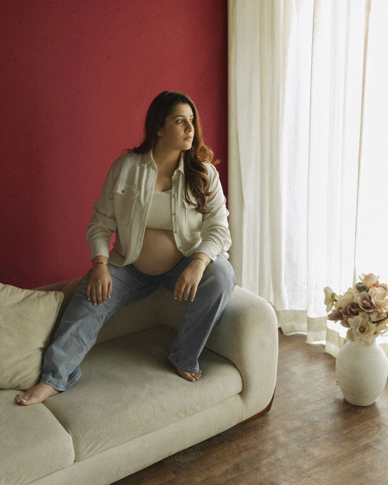 Pregnant woman in white jacket and jeans sits on cream sofa against red wall, gazing thoughtfully toward sheer curtains.
