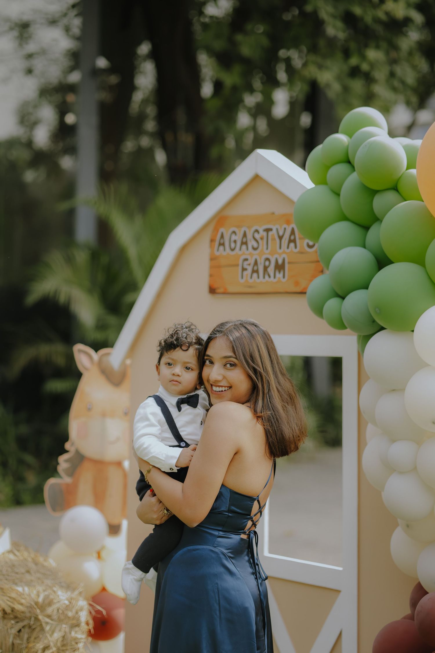 Woman in blue dress holds baby boy at Agastya's Farm birthday party with green and white balloon decorations.
