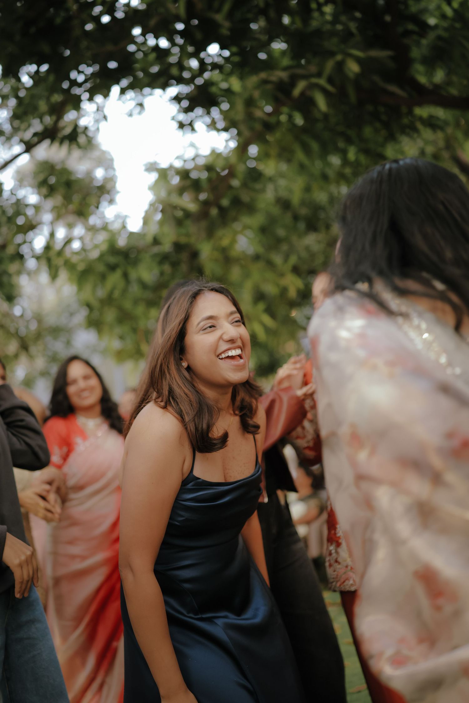 A young woman in a black satin dress laughs joyfully at an outdoor gathering with guests in colorful attire.