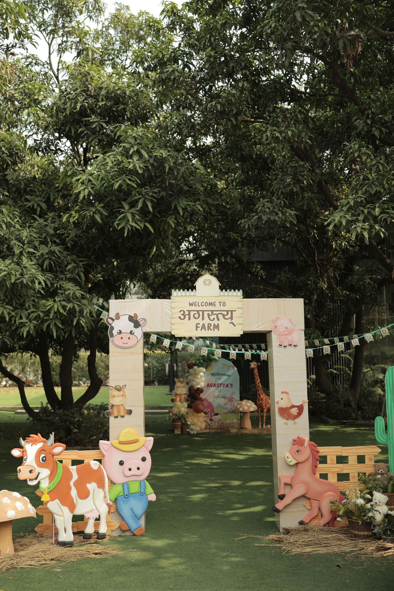 Farm-themed birthday party entrance arch with cartoon animal cutouts, Hindi text, and lush green trees backdrop.