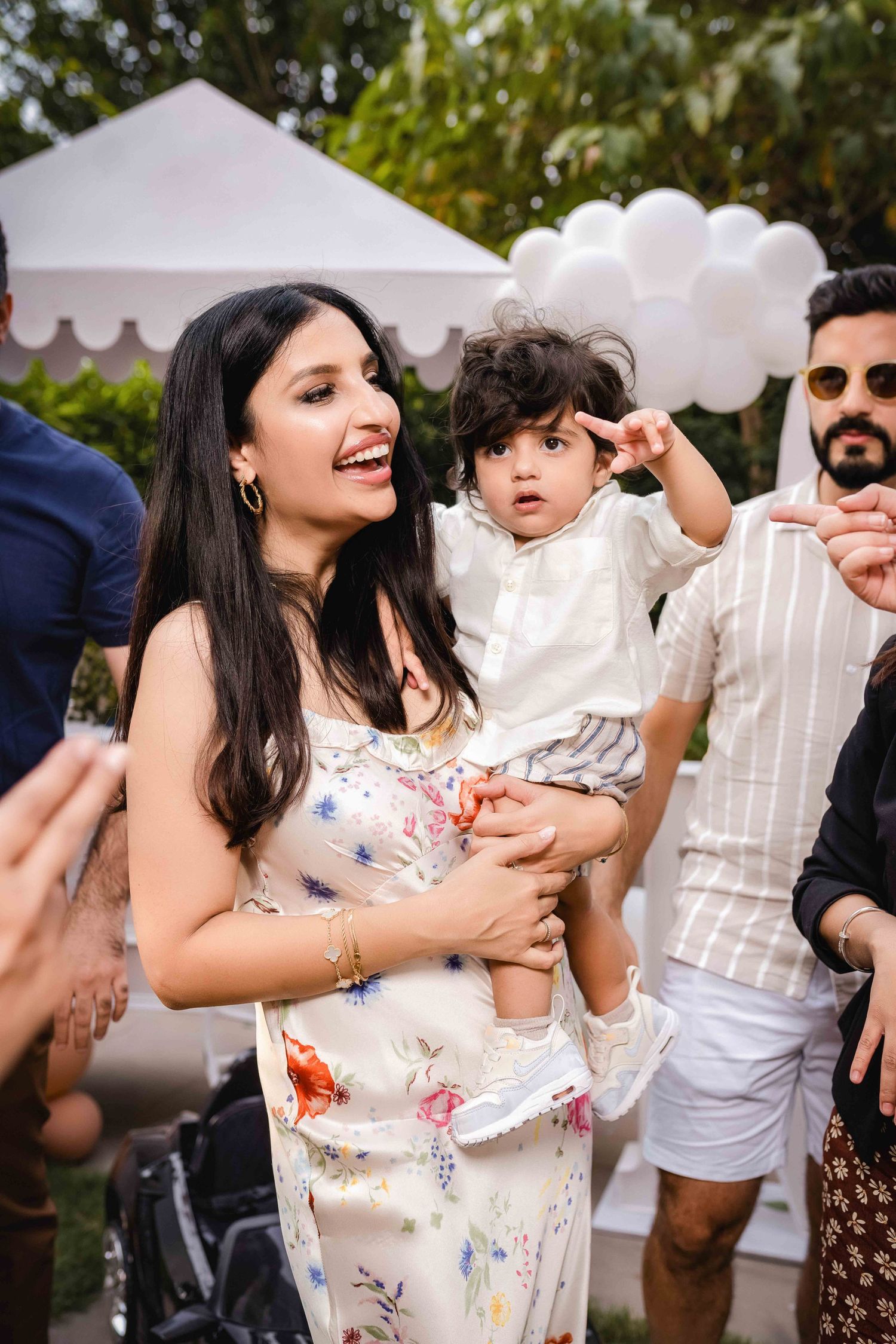 Woman in floral dress holding a toddler at an outdoor party with white balloons and guests in background.