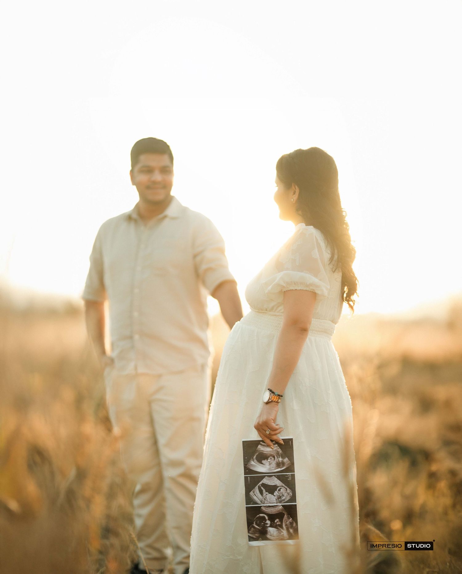 Couple holding ultrasound photos in golden field at sunset, woman in white dress, maternity announcement shoot.