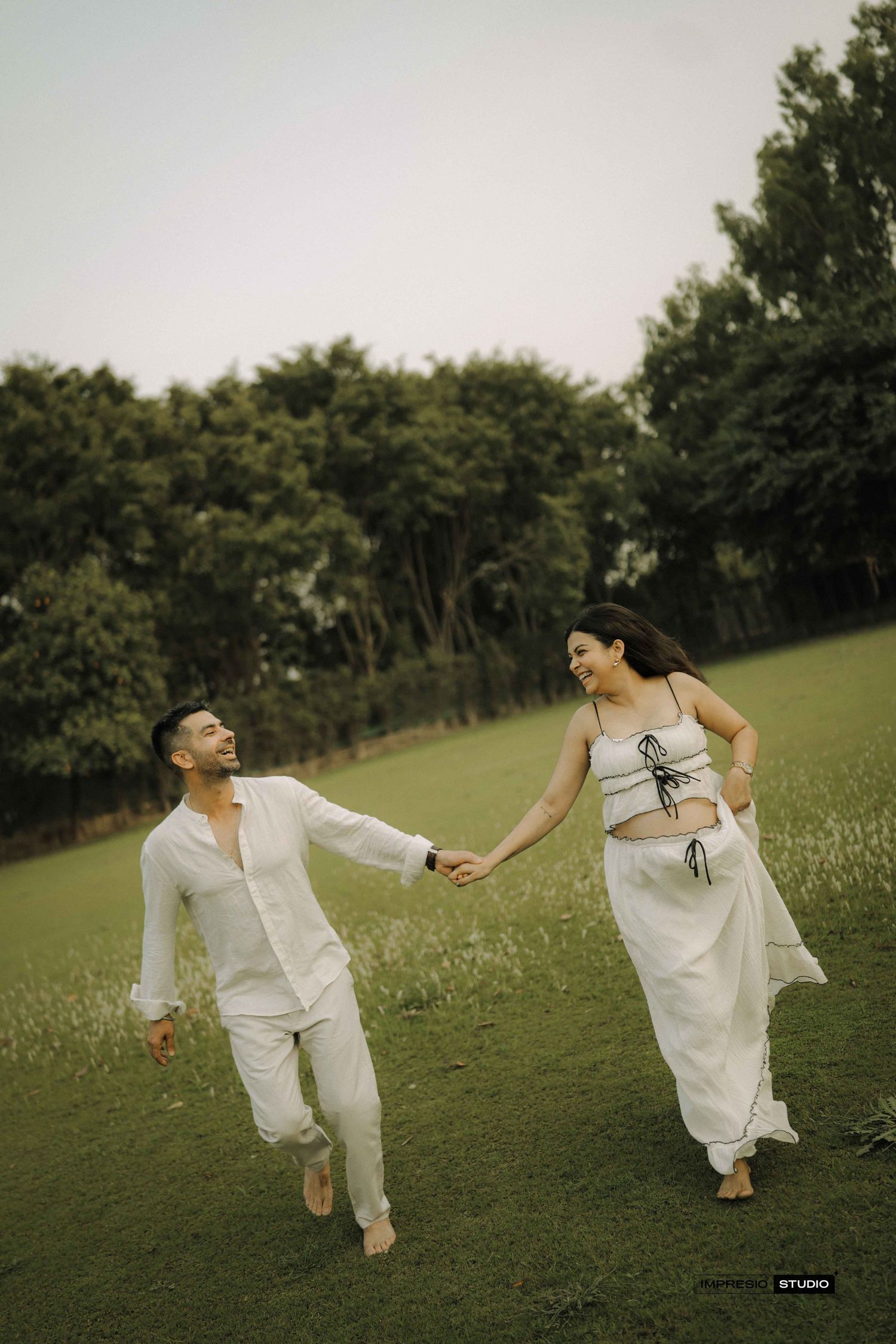 Happy couple in matching white outfits holding hands while walking barefoot on green meadow.