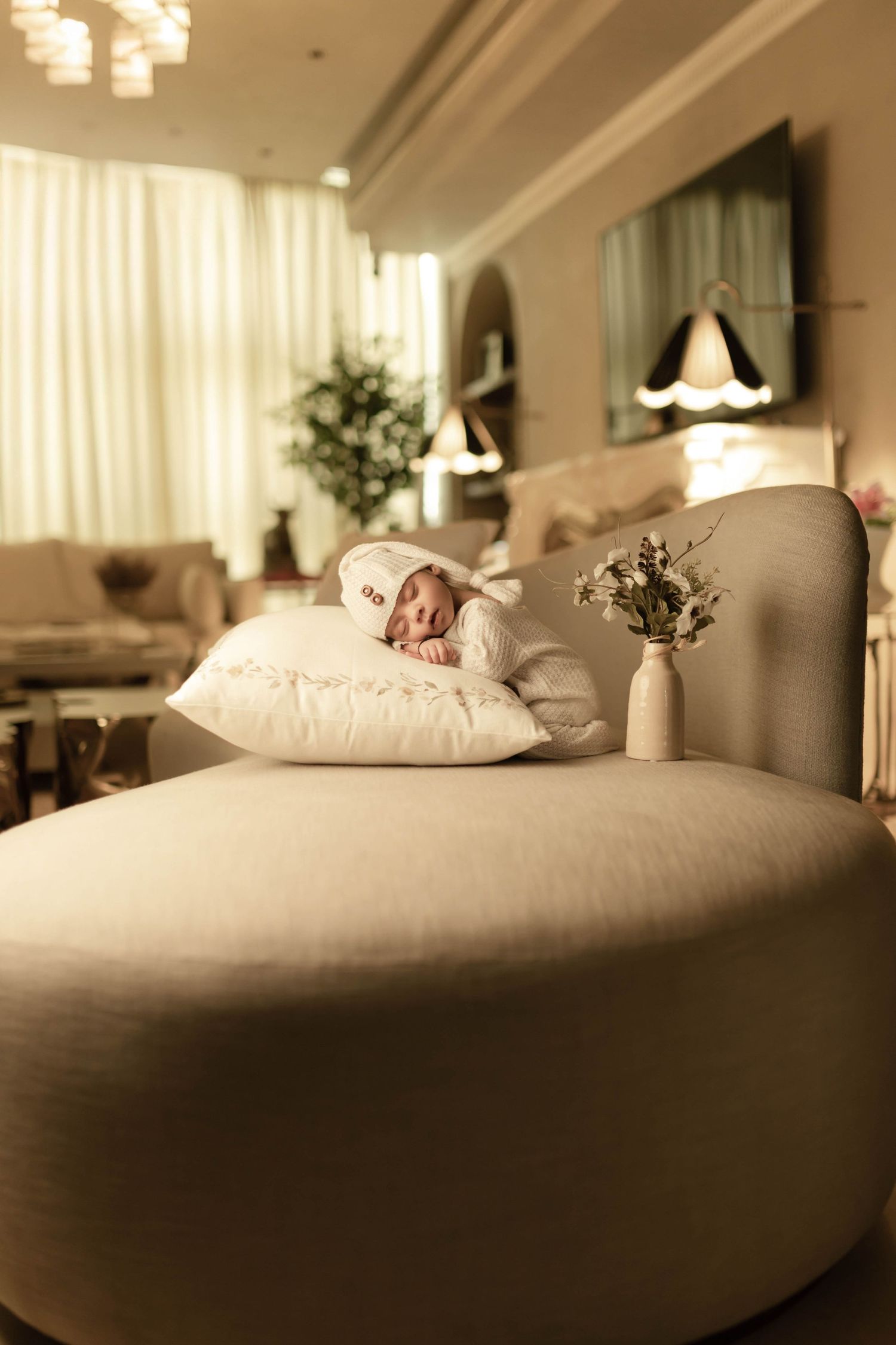 A newborn baby in a white outfit sleeps peacefully on a pillow atop a beige chaise lounge in an elegant room.