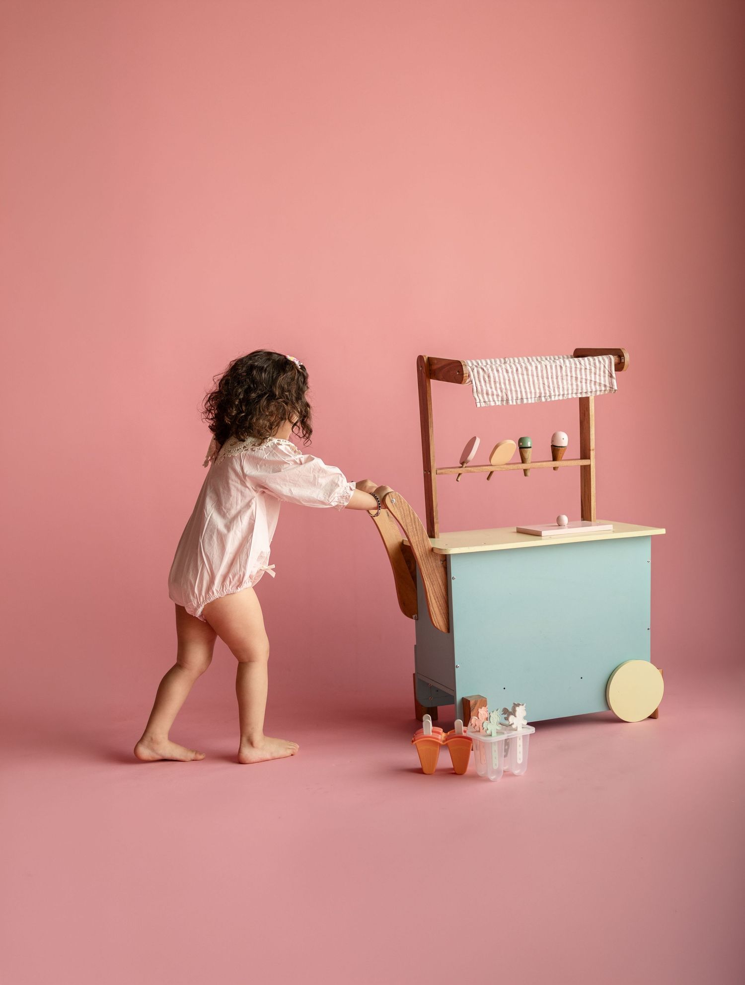 Young child playing with a pastel blue wooden toy ice cream cart against a pink background.