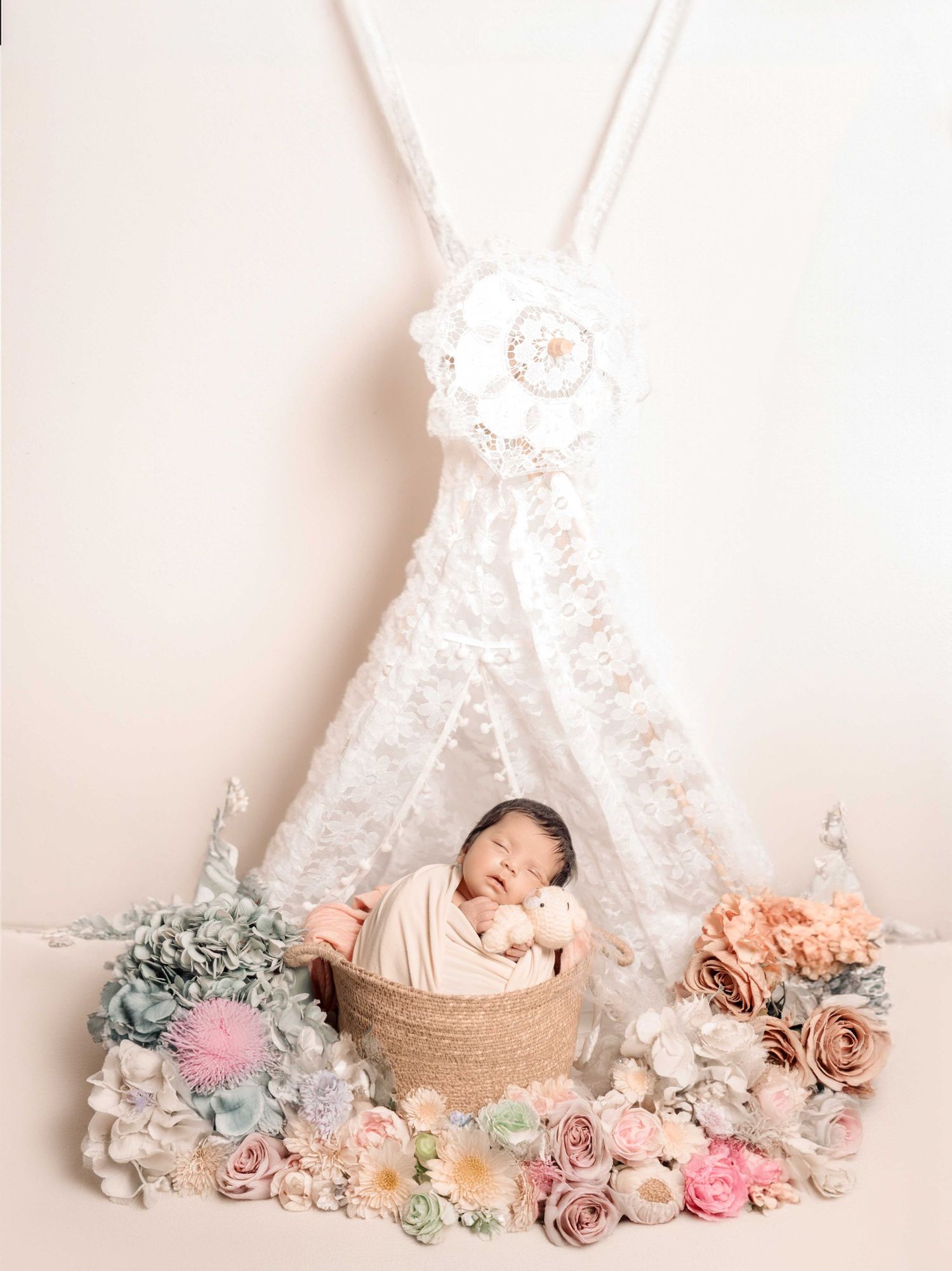 Newborn baby sleeping in a basket surrounded by pastel flowers under a white lace teepee in a soft studio setting.