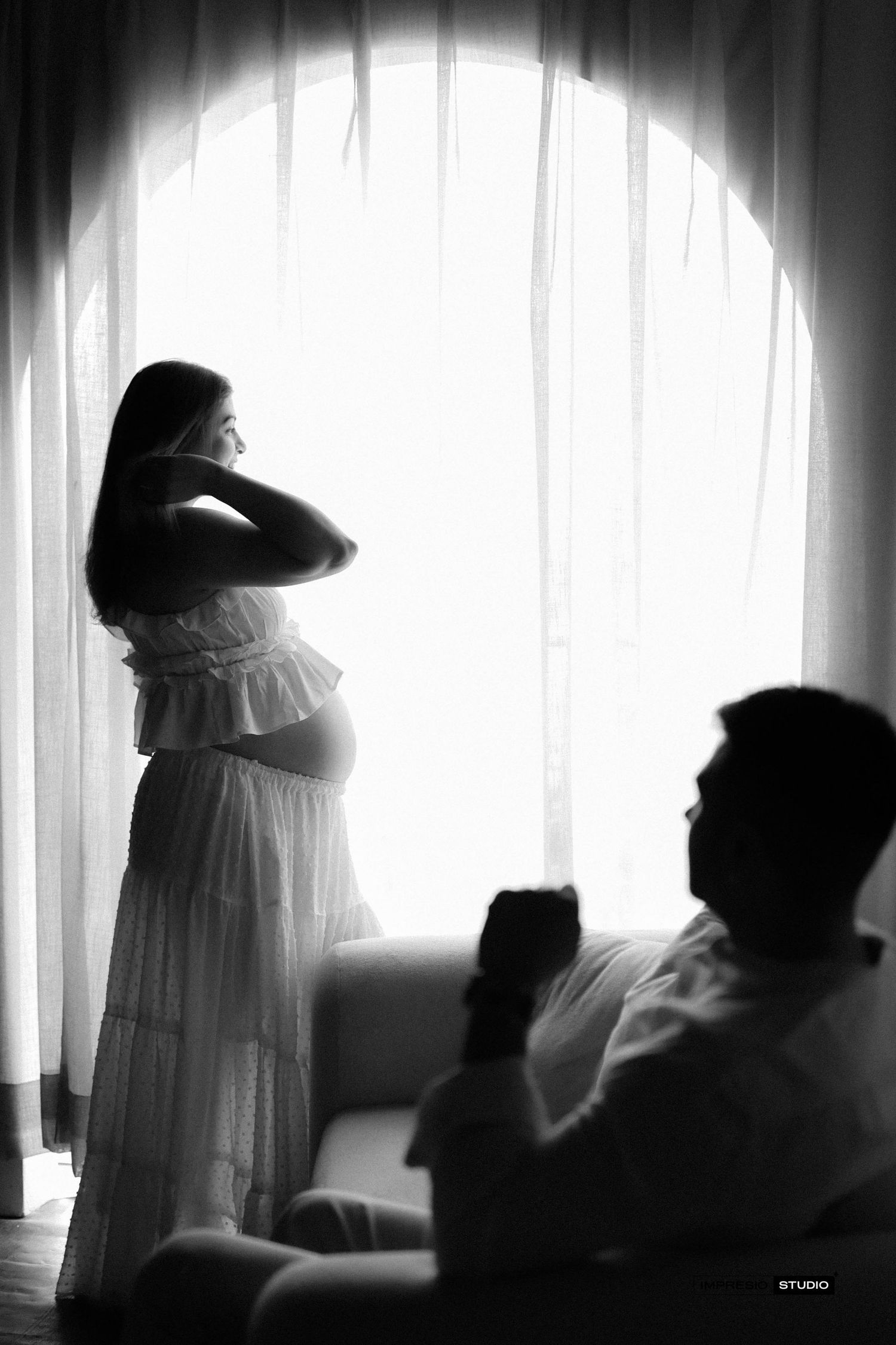Elegant black and white maternity photo of a pregnant woman in a white dress standing by an arched window with soft backlight.