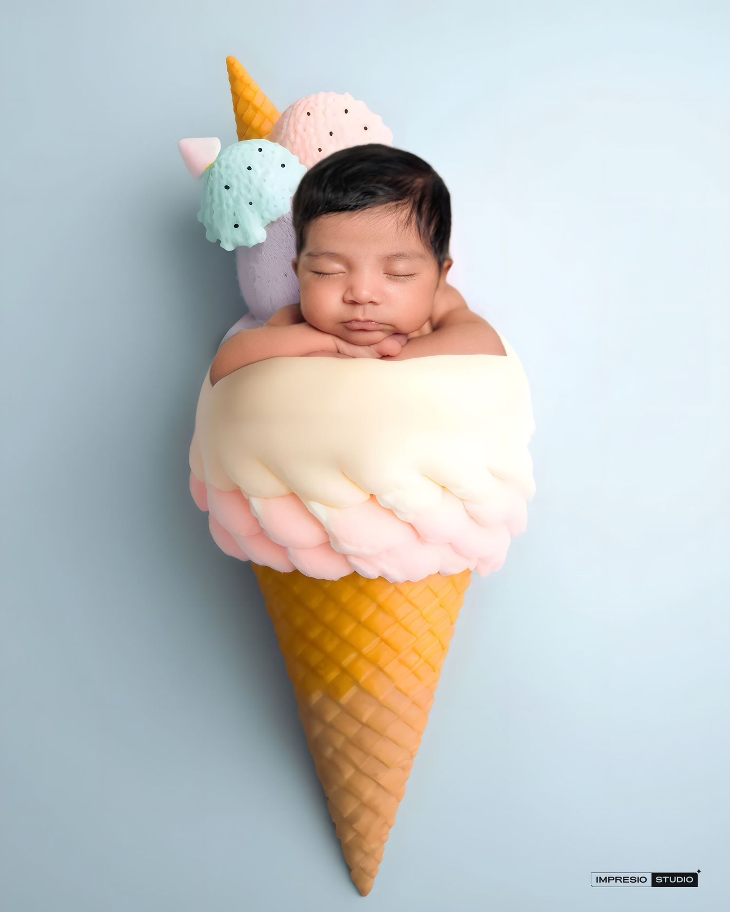 Newborn baby sleeping peacefully inside a large decorative ice cream cone prop with pastel scoops on blue background.