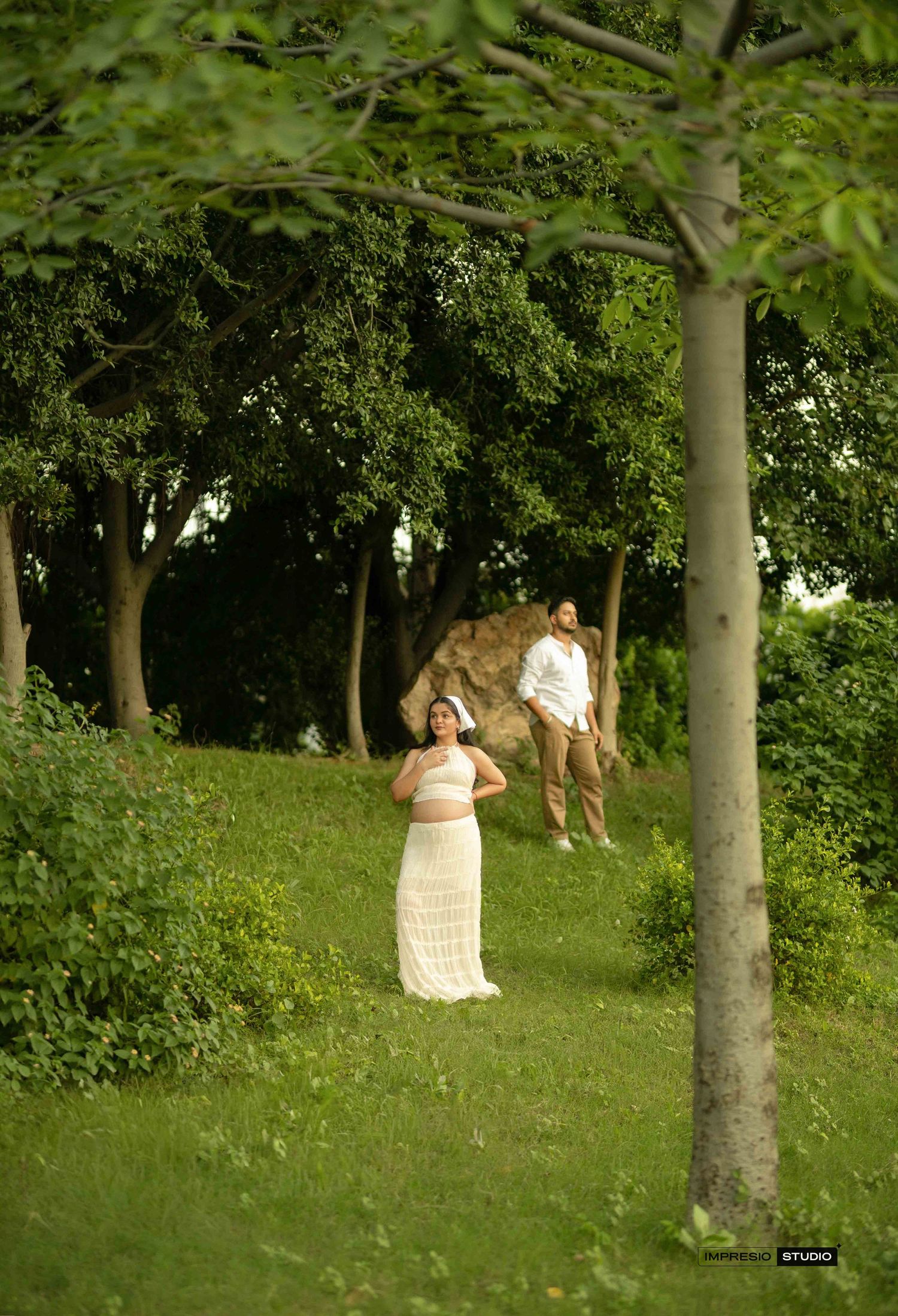 A couple in white attire poses romantically in a lush green park surrounded by tall trees.