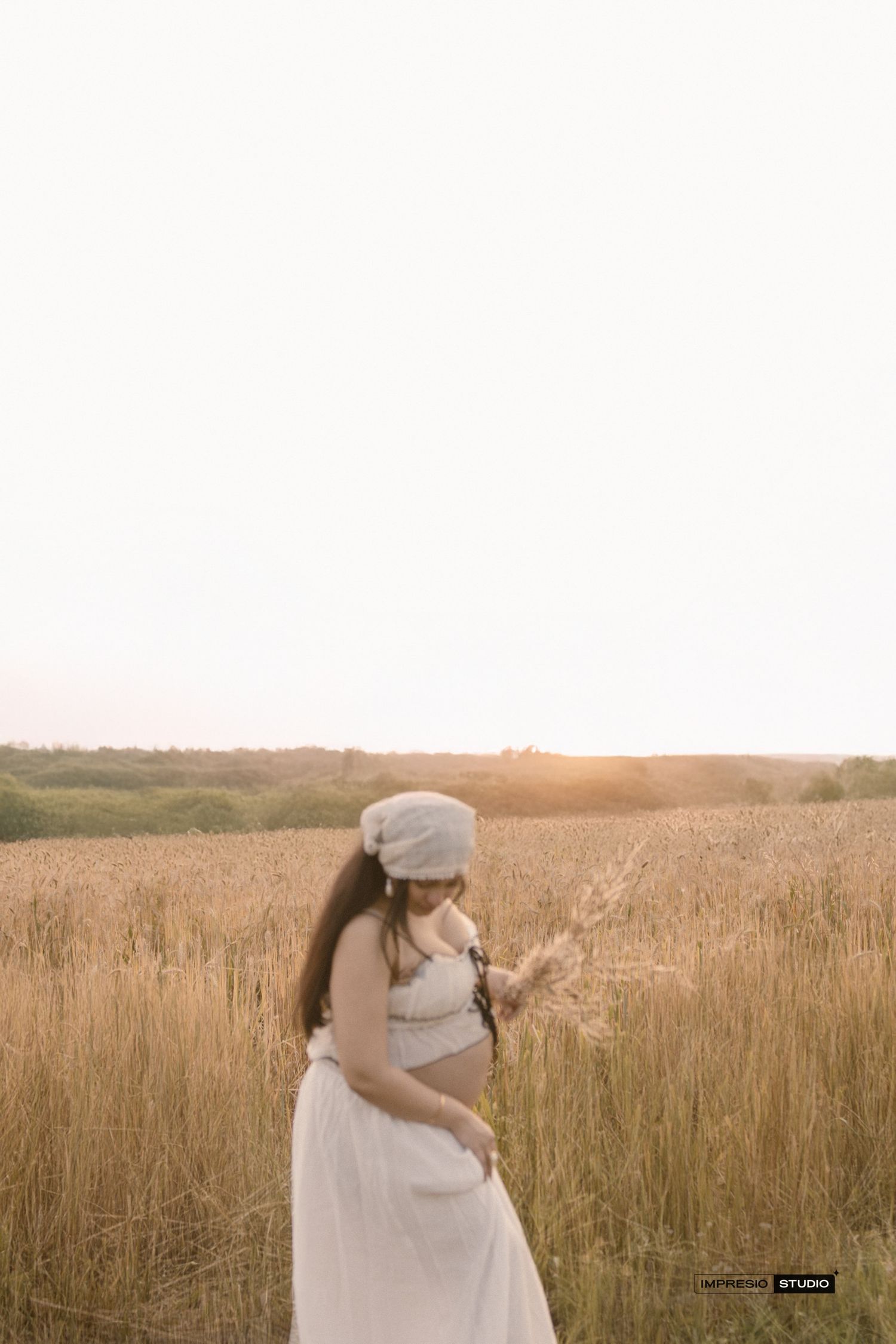 Pregnant woman in white skirt and beanie holding wheat stalks in golden field at sunset.