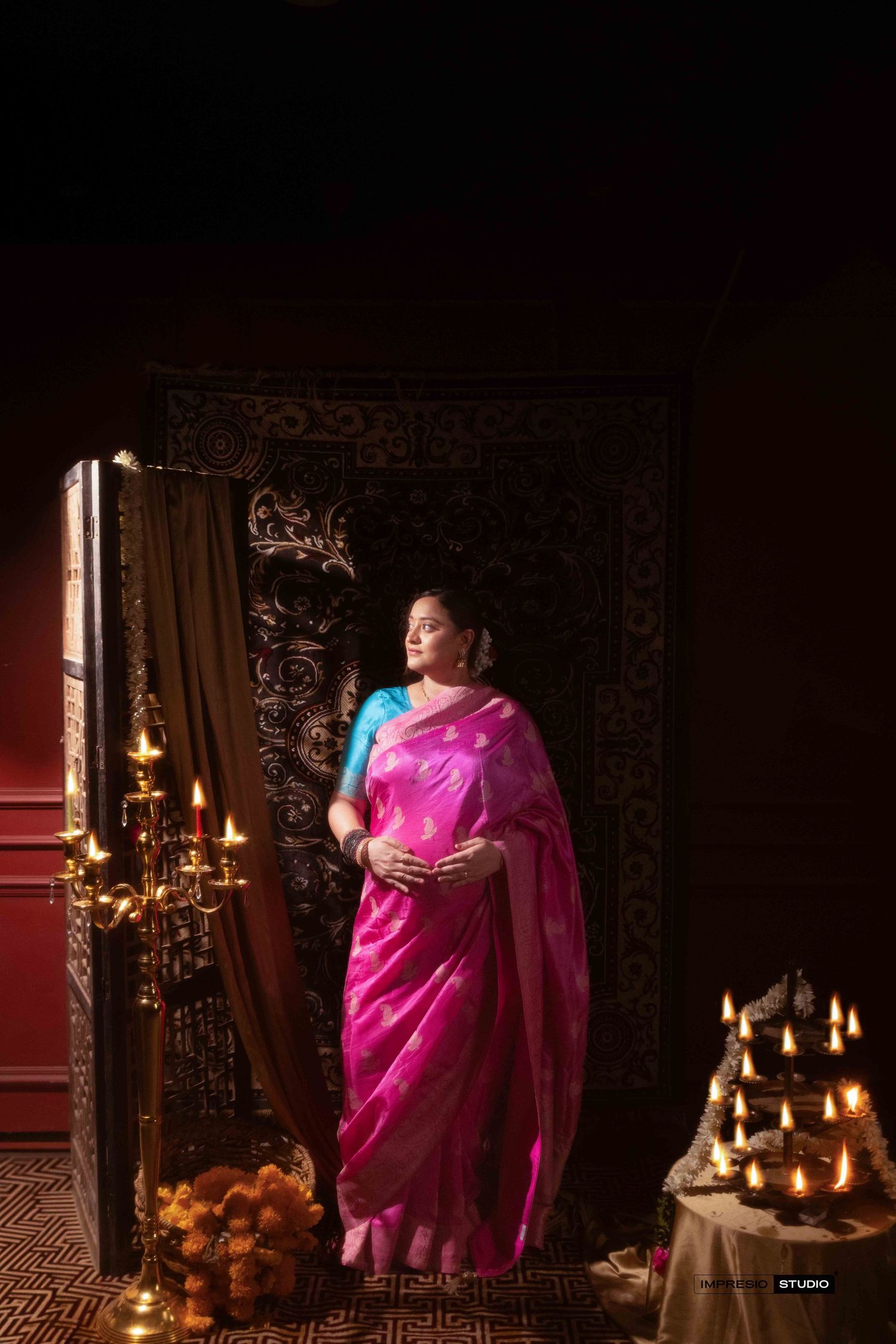 Pregnant woman in pink silk saree with blue blouse poses gracefully among lit diyas and candelabra in dark studio.