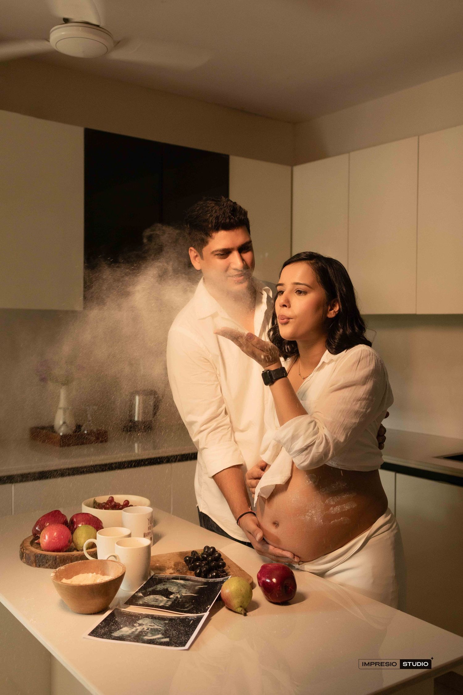 Pregnant couple playfully blowing flour in a modern kitchen, surrounded by fresh fruits and ultrasound photos.
