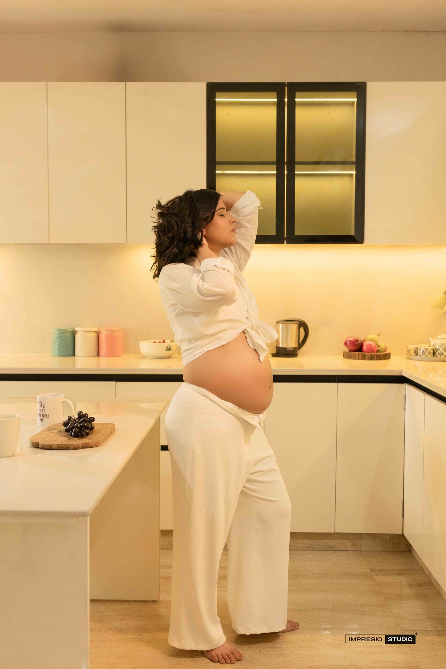 Pregnant woman in white outfit standing in modern kitchen showing baby bump.