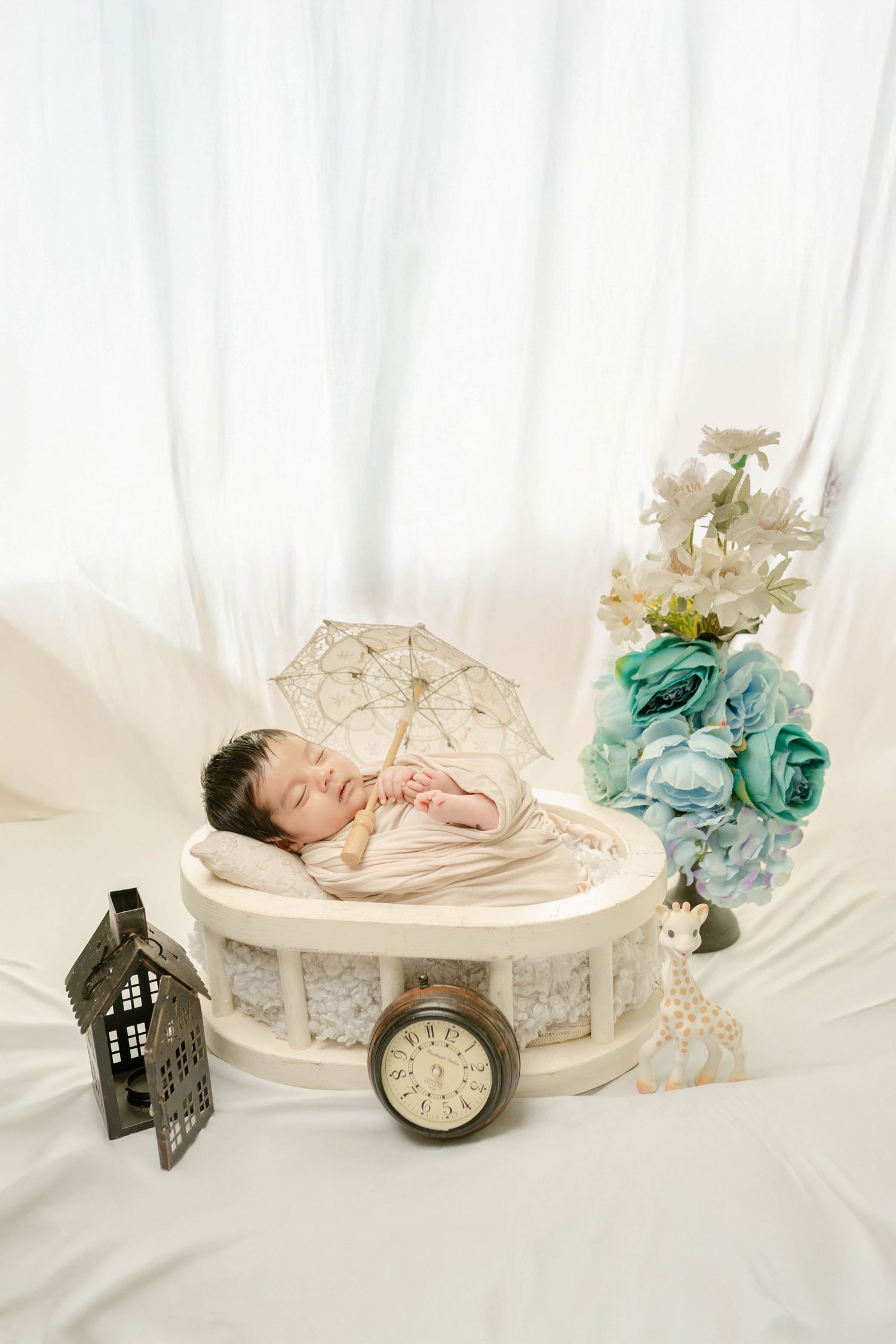 Newborn baby sleeping in a round white bed with a lace umbrella, flowers, clock, and decorative props on white fabric.