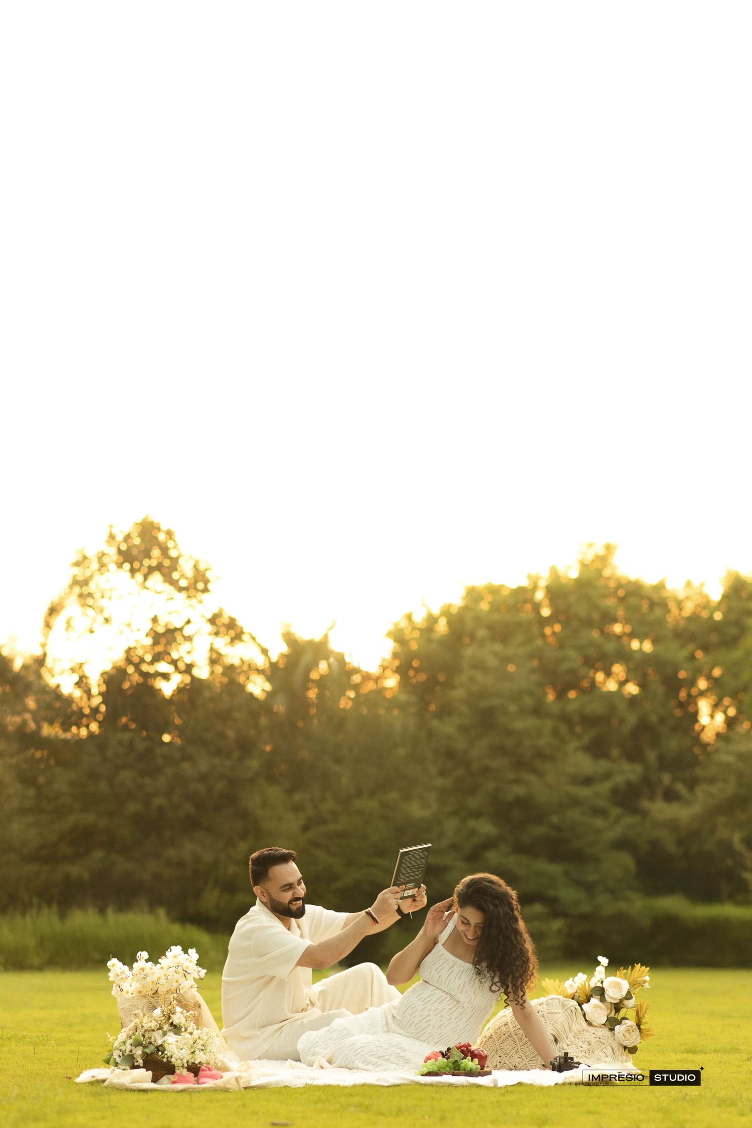 Couple in white attire enjoying a romantic outdoor picnic with flowers at golden hour in a lush green park.