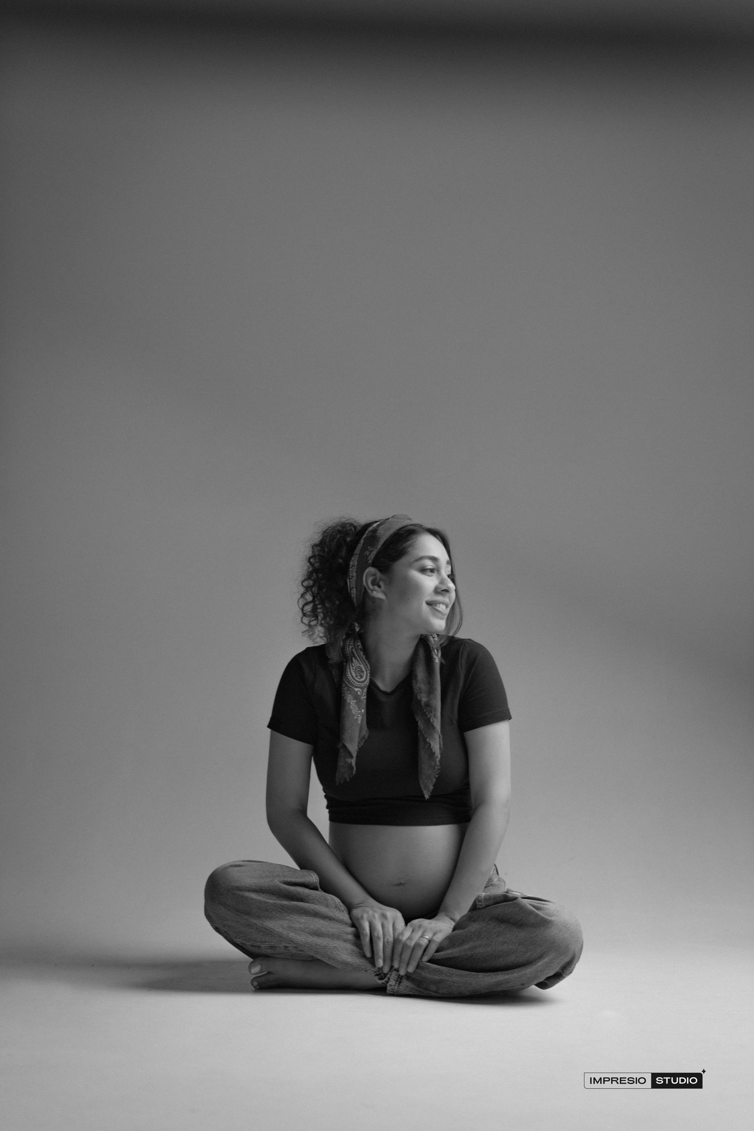 Black and white photo of a pregnant woman sitting cross-legged on studio floor, smiling, wearing crop top and loose pants.